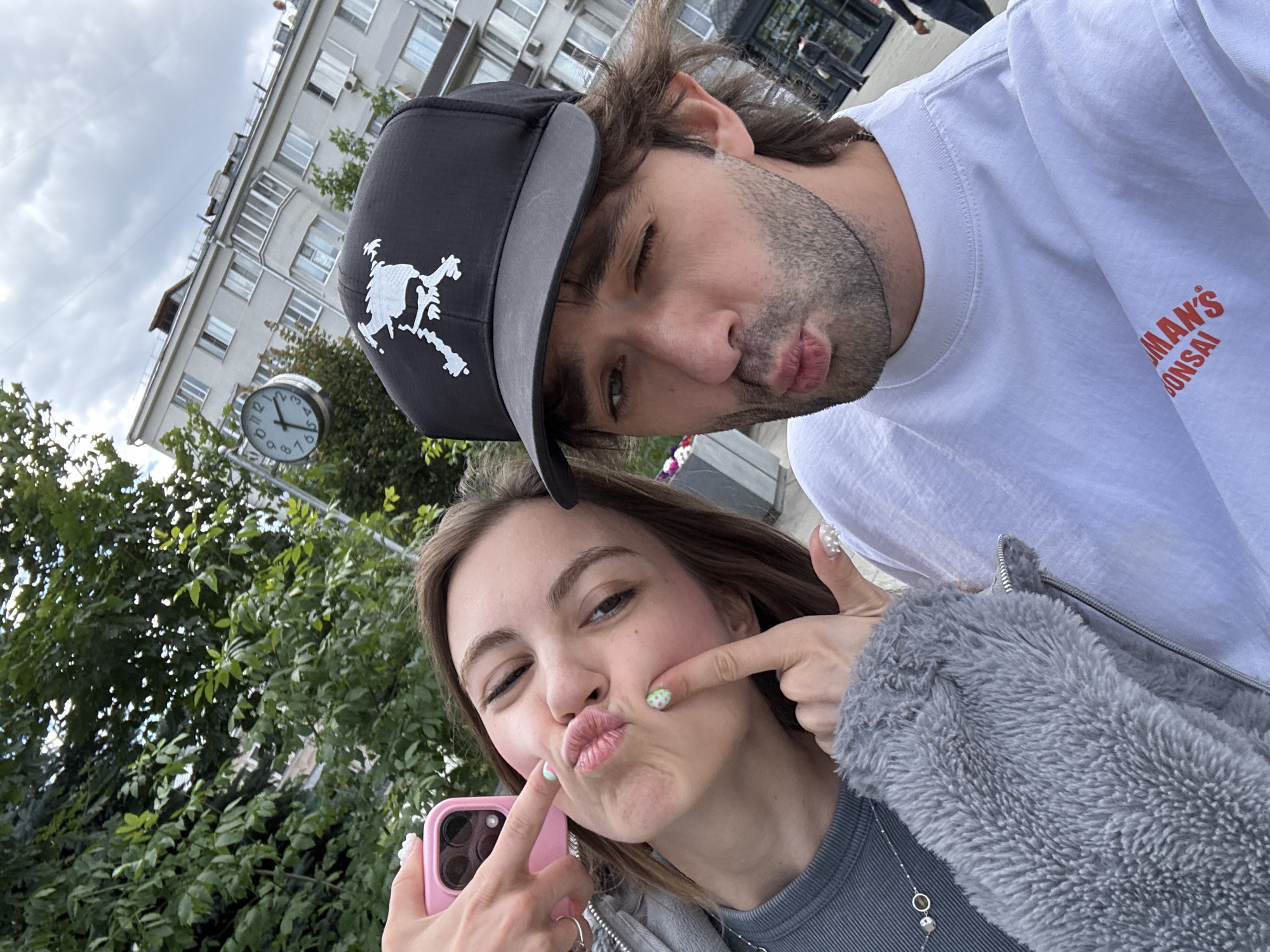 Two people, a man and a woman, are taking a selfie outdoors. The man is wearing a black baseball cap with a white graphic of what appears to be a skeleton in a running pose and a white t-shirt with orange text that reads "HUMAN'S BONSAI". He is winking and puckering his lips. The woman is to his left, also puckering her lips and playfully poking her cheeks with her fingers. She is holding a pink smartphone in her left hand. She is wearing a grey fuzzy jacket over a grey top. Her fingernails are decorated with white and green nail art.
In the background, there is a building with multiple windows and balconies, and lush green trees. A clock is visible on a pole, showing the time as approximately 1:30. The sky is overcast, suggesting a cloudy day. The scene appears to be in an urban setting, possibly Moscow, Russia, given the context. The overall mood is playful and casual.