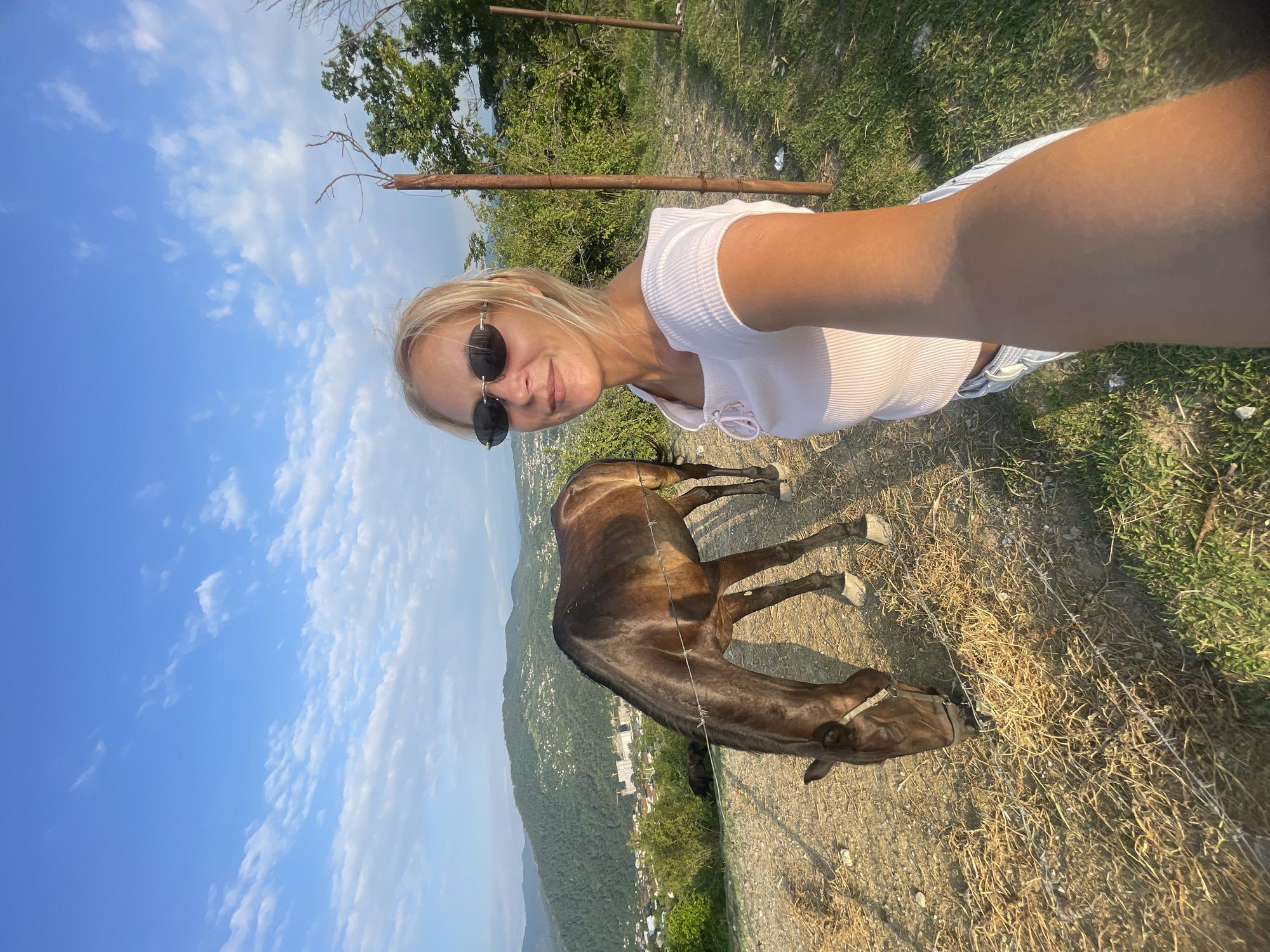 A blonde woman in a white crop top and sunglasses takes a selfie in front of a brown horse grazing on dry grass. The horse is wearing a bridle. The woman's arm is extended, holding the camera. In the background, rolling green hills dotted with buildings descend towards a distant cityscape. The sky is blue with scattered white clouds. The location appears to be an elevated area overlooking the city of Baranovka, Russia. The scene is bathed in the warm light of what seems to be late afternoon or early evening.