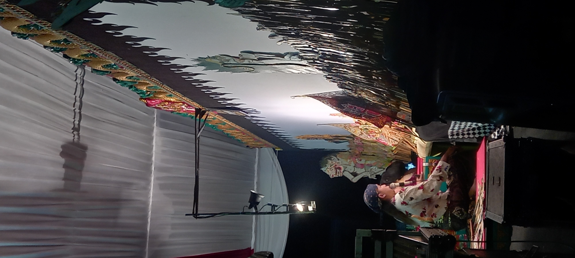 The image shows a shadow puppet performance, known as Wayang Kulit, in Wanareja, Indonesia. A performer, dressed in traditional Indonesian clothing including a patterned headscarf and a floral shirt, is seated on the floor, facing a white screen. The performer is holding a microphone and appears to be narrating or singing. Behind the performer, various shadow puppets are arranged, some in the process of being manipulated or displayed against the screen, casting shadows. The screen is adorned with elaborate, colorful decorations along its top edge, featuring mythical figures and intricate patterns. A spotlight is directed towards the screen, illuminating the puppets and creating the shadows. The overall setting suggests a cultural event or performance taking place indoors, possibly under a tent or temporary structure, as indicated by the draped white fabric forming the ceiling. The presence of a microphone and the illuminated screen are characteristic of a Wayang Kulit performance.