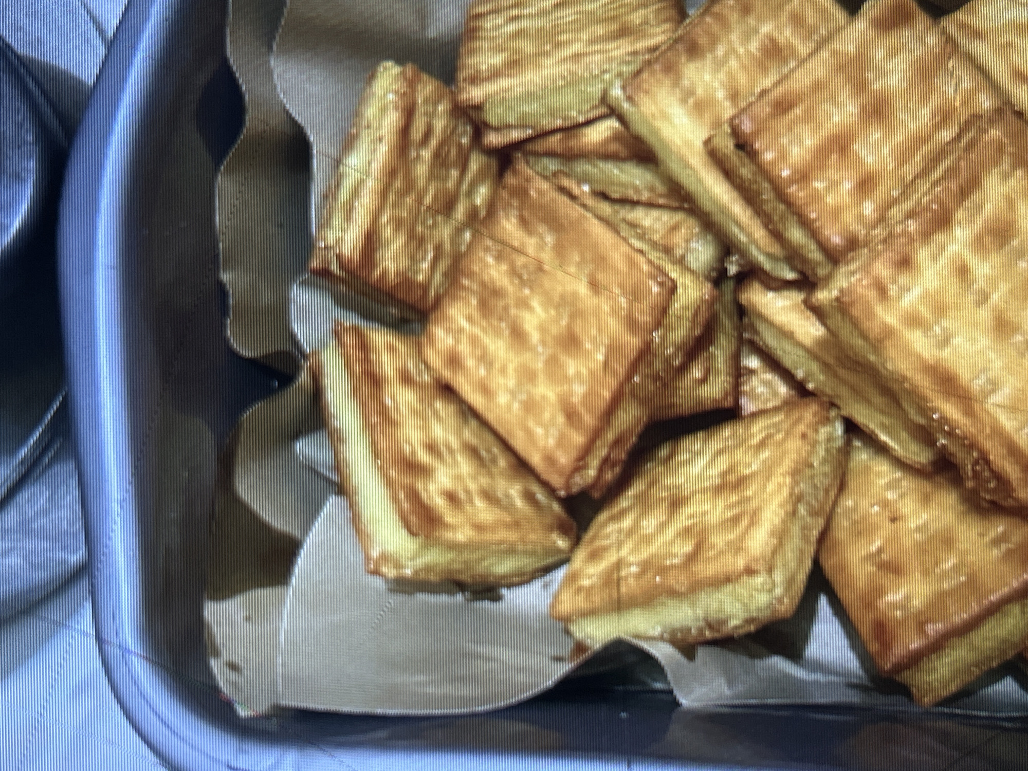 A close-up, overhead shot shows a metal container filled with square-shaped biscuits. The biscuits are golden brown and appear to have a textured surface. They are layered and stacked within the container, which is lined with brown paper. The image is slightly out of focus and exhibits digital noise, with horizontal lines present throughout. The context suggests these biscuits are likely a food item, possibly being prepared or served. There is no identifiable location within the city of Depok, Indonesia, visible in the image.