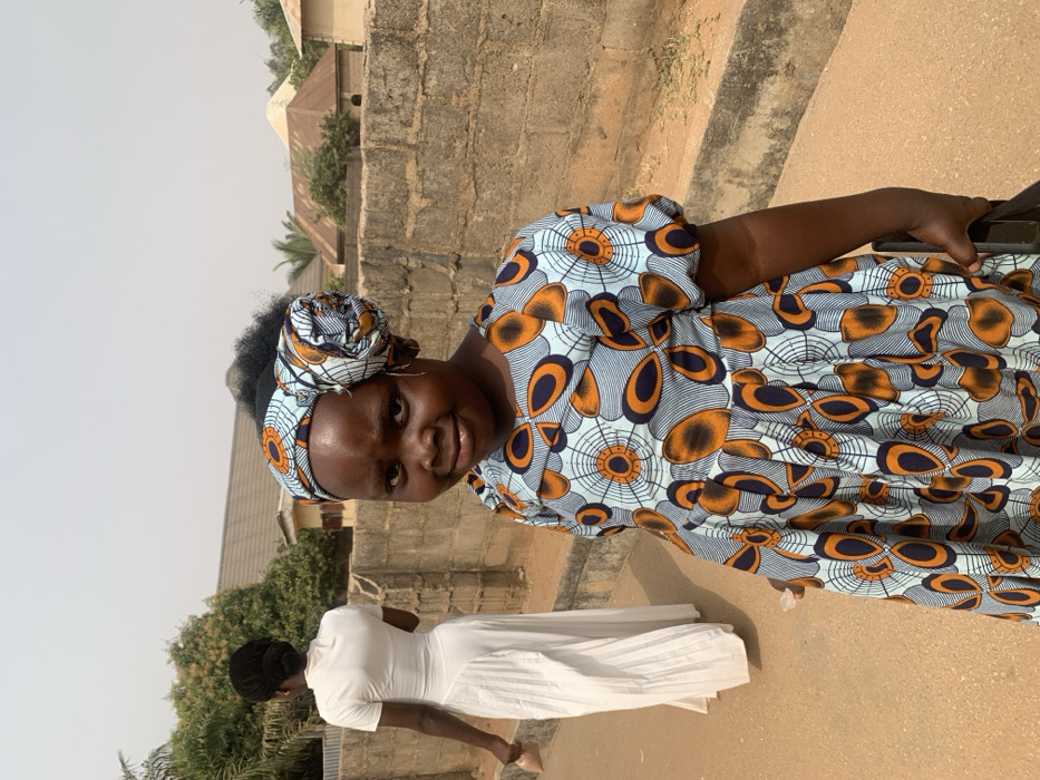 An outdoor scene in Garaku, Nigeria, features two females. In the foreground, a young female with dark skin wears a light blue dress patterned with orange and dark blue concentric circles and elongated shapes, featuring puffed sleeves and a pleated skirt. Her head is adorned with a matching fabric headwrap styled into a prominent roll on the right side, with dark, curly hair visible. She displays a slight smile and wears small hoop earrings. Her left hand is partially visible, holding a dark object. In the mid-ground, an adult female with dark skin, wearing a long, white, high-collared dress with short sleeves and a pleated section, walks away from the camera, holding a light-colored object in her right hand. Her hair is pulled back into a bun. The ground consists of unpaved, light brown earth. A concrete block wall runs along the right side of the frame. Beyond the wall, various structures are visible, including one with a light-colored, possibly conical roof, and scattered green foliage under a clear, light blue sky.