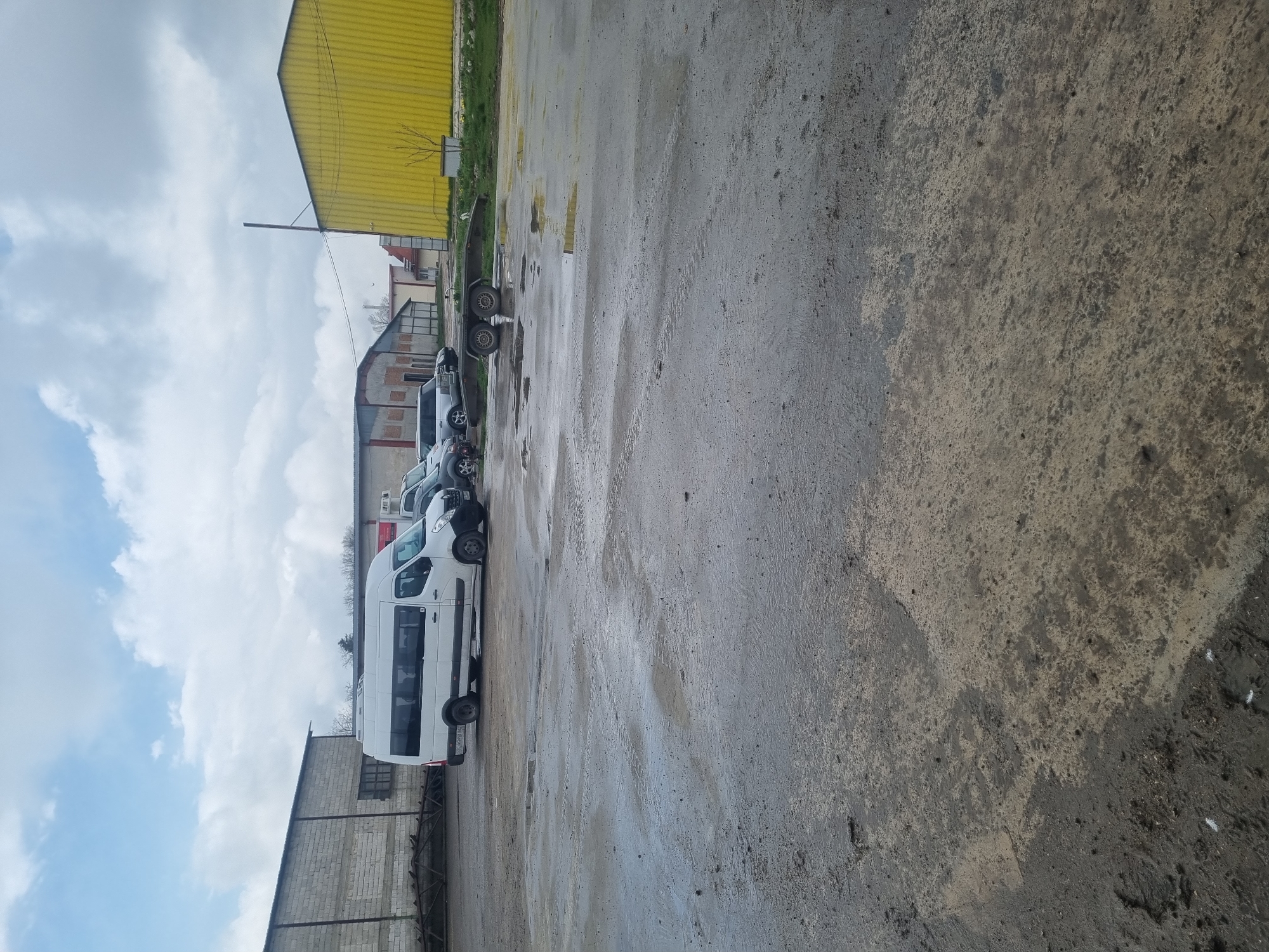The image shows a wet, paved lot in Ovcharovo, Bulgaria, with several white vans parked in a row. A yellow industrial building is visible in the background, along with other buildings. The sky is cloudy. The ground surface appears to be a mix of concrete and asphalt with water puddles.
