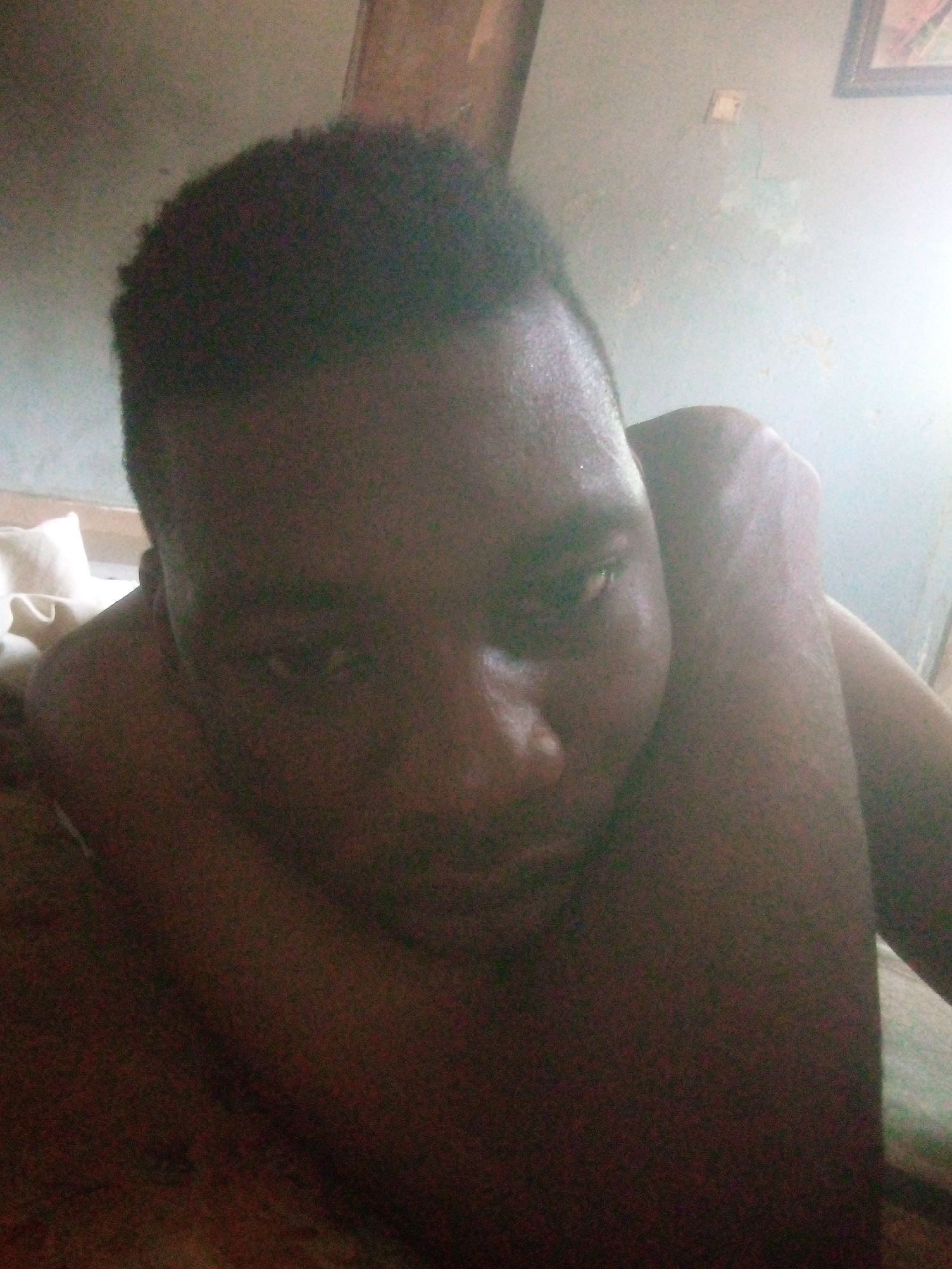 A close-up, low-angle shot shows a Black man lying down. The background consists of a wall with a framed picture and what appears to be an electrical outlet. A pillow is visible to the left. The location is given as the city of Unknown, Unknown.