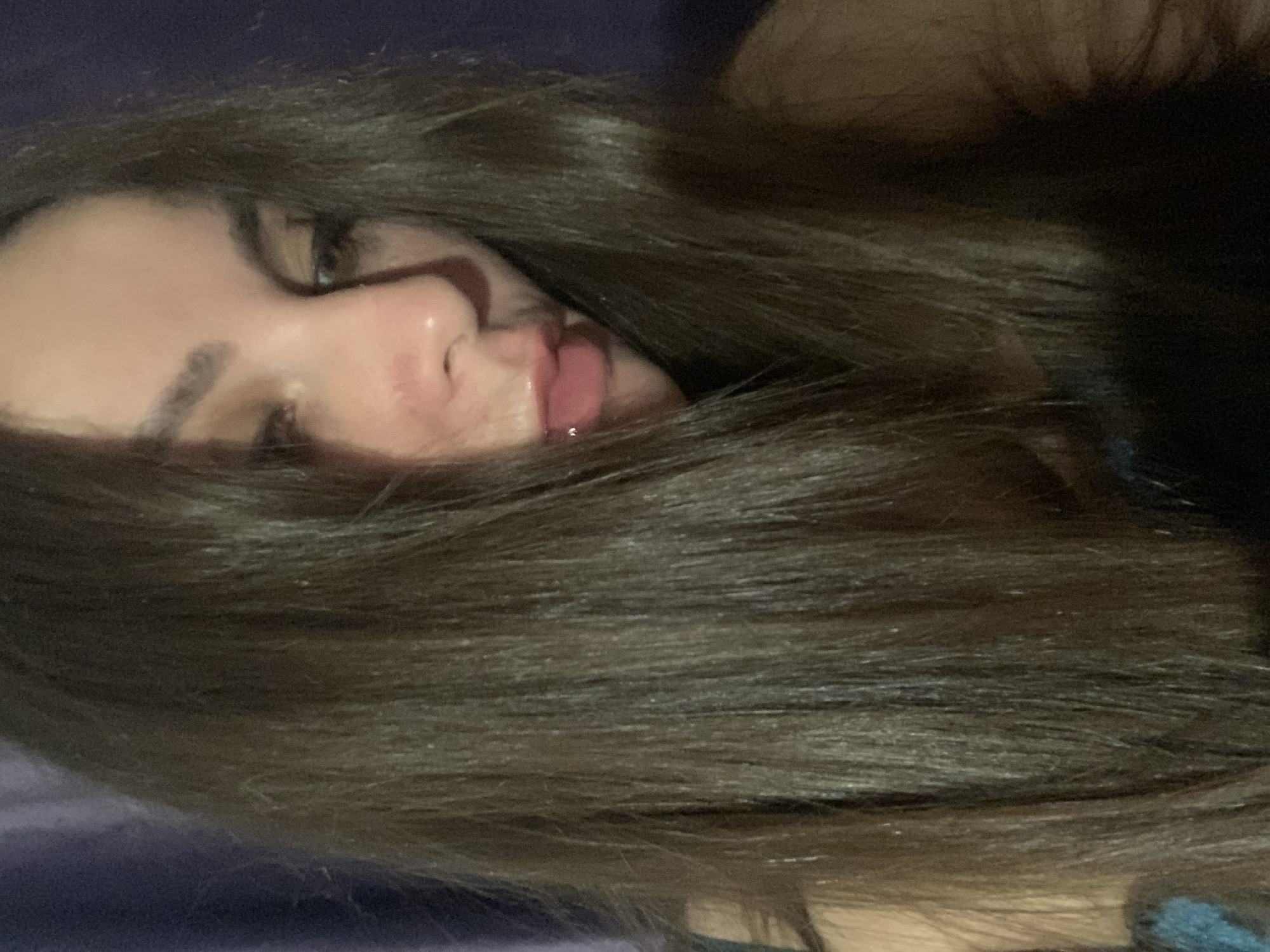 A young woman with long, dark brown hair is captured in a close-up, downward-facing shot against a dark purple background. Her face is partially obscured by her hair, with her nose and lips visible. Her lips are pink and appear to be slightly parted, with a subtle gloss. Her eyebrows are dark and well-defined. The lighting is dim, casting soft shadows across her features. The image appears to be a selfie taken indoors, with no discernible urban features.