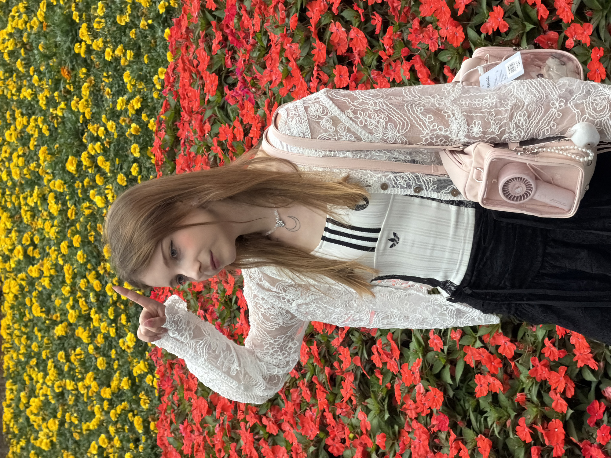 A young woman stands in front of a vibrant floral display. The background is filled with a dense arrangement of red and yellow flowers. The woman has long brown hair and is wearing a white lace cardigan over a black and white striped top with Adidas and a Trefoil logo. She is also wearing a black skirt and a light pink shoulder bag that contains a pink hair dryer in a clear compartment and a fuzzy white charm. She is holding her right hand up to her forehead and pointing her index finger towards the sky. The setting appears to be an outdoor garden or park, possibly in the city of Yuzhong District, China, given the context.