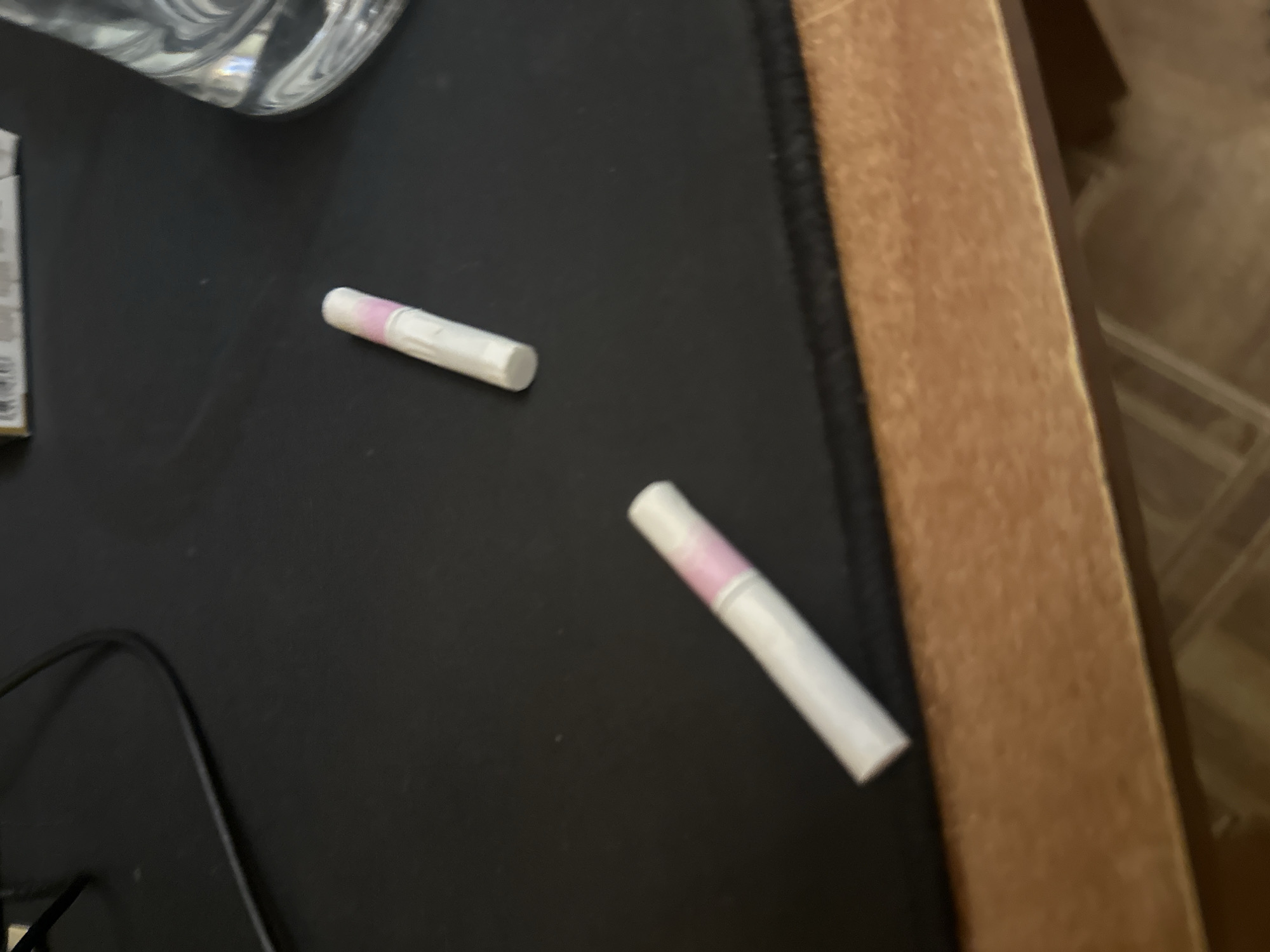 The image shows two white and pink cylindrical objects, possibly swabs or small tubes, lying on a black mousepad next to a wooden desk or table. Part of a clear glass object is visible in the upper left corner, and a black cable is also present. The setting appears to be an indoor environment, possibly a home office or study. The location in the city is unknown.