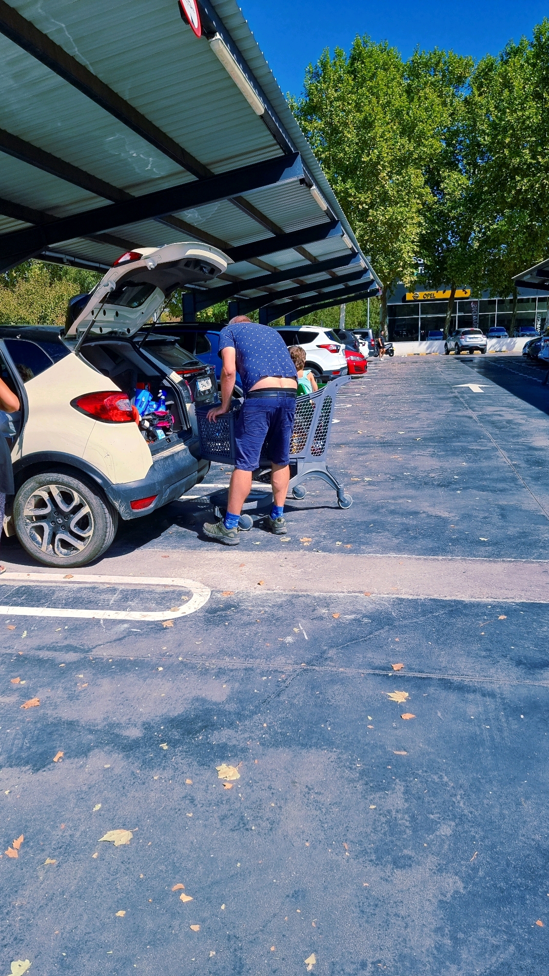 The image depicts a man loading items into the open trunk of a light-colored car parked in an outdoor parking lot covered by a dark-colored roof structure. The man is wearing a blue patterned short-sleeved shirt and blue shorts, with green shoes and blue socks. He is bent over, reaching into a shopping cart that is positioned next to the car's trunk. Inside the trunk, various items, including blue bottles, are visible. Another person, partially visible on the left, appears to be standing near the car. In the background, several other cars are parked, and a building with the "OPEL" logo is visible. Lush green trees are present behind the building. The parking lot surface is dark asphalt with some scattered fallen leaves. The scene appears to be in a commercial area, likely a shopping center parking lot. There is no specific indication of the location being Olot, Spain.