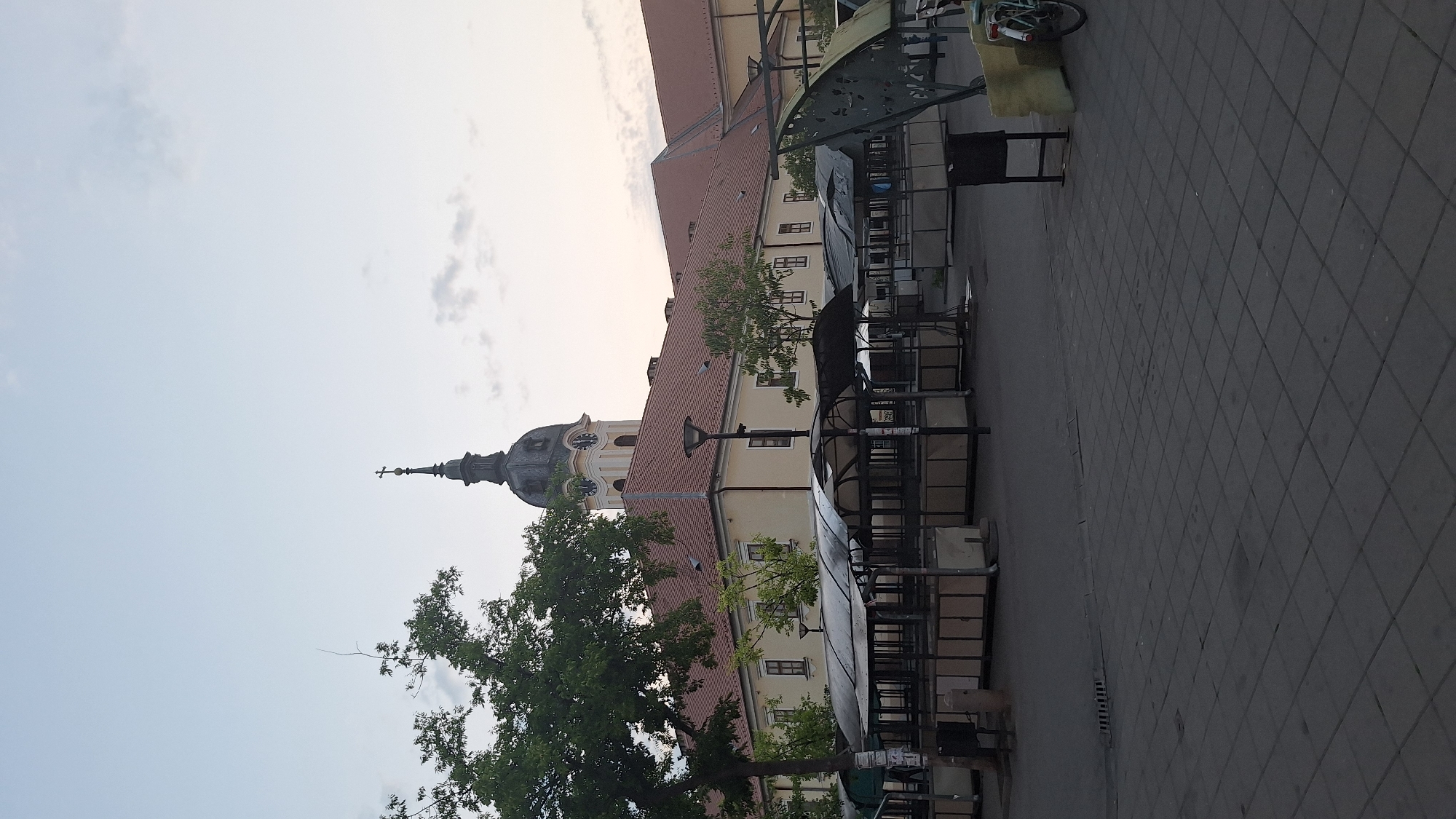 In the city of Sombor, Serbia, this photograph captures an outdoor market area with a large, yellow-colored building in the background. The building features a red tiled roof and a prominent clock tower with a cross at its peak. In front of the building are several empty market stalls, covered with a dark fabric, suggesting the market is not currently active. The ground is paved with dark grey square tiles. To the right of the stalls, a bicycle is visible, leaning against a yellow object. The sky is clear with a few wispy clouds, and the overall lighting suggests it is either early morning or late evening.