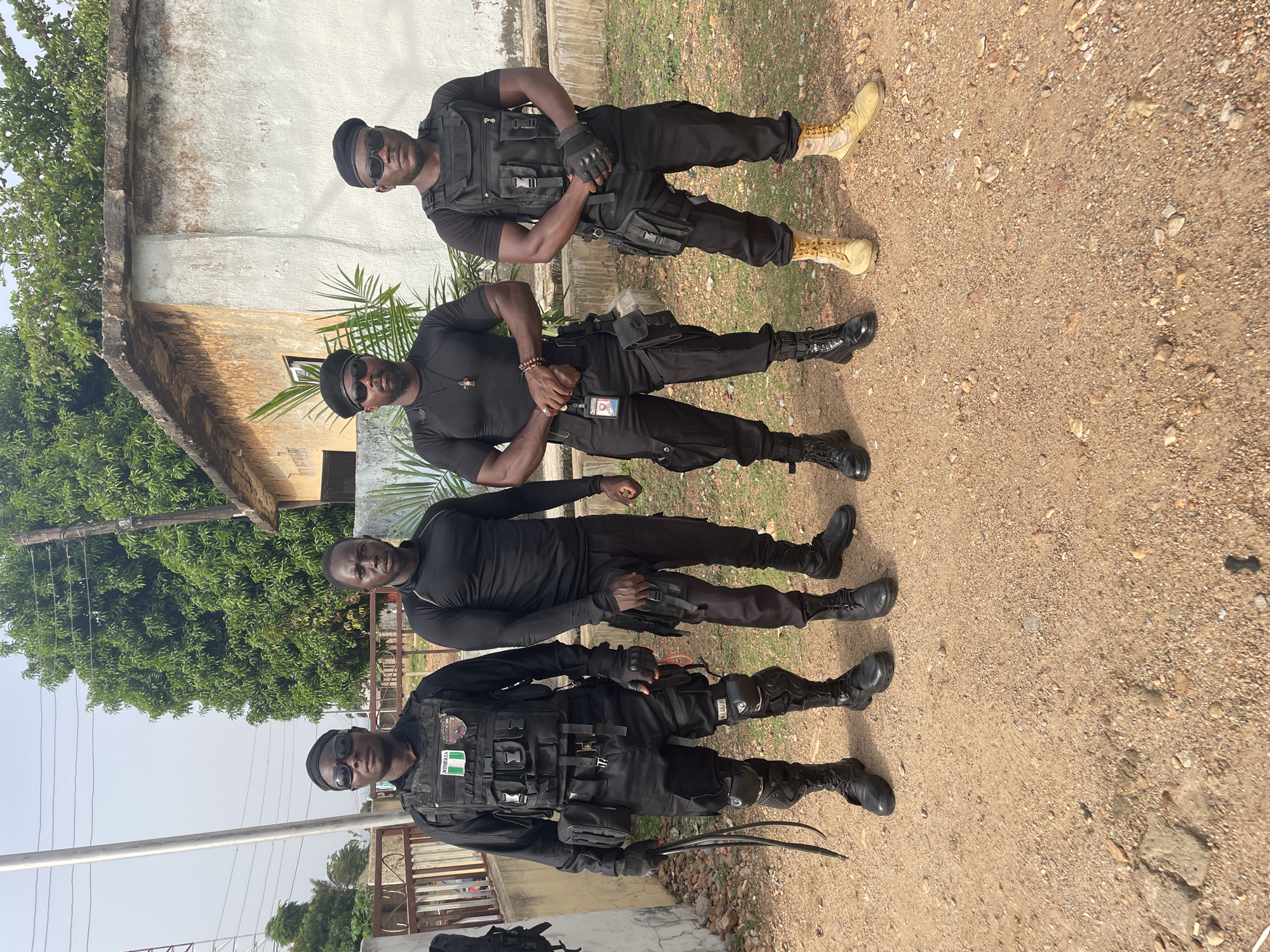 Four men are posing in front of a building and a dirt yard. The men are all wearing black clothing. The man in the bottom of the image is wearing a tactical vest with a Nigerian flag patch. The man second from the top is wearing khaki boots. The location may be Akwanga, Nigeria.