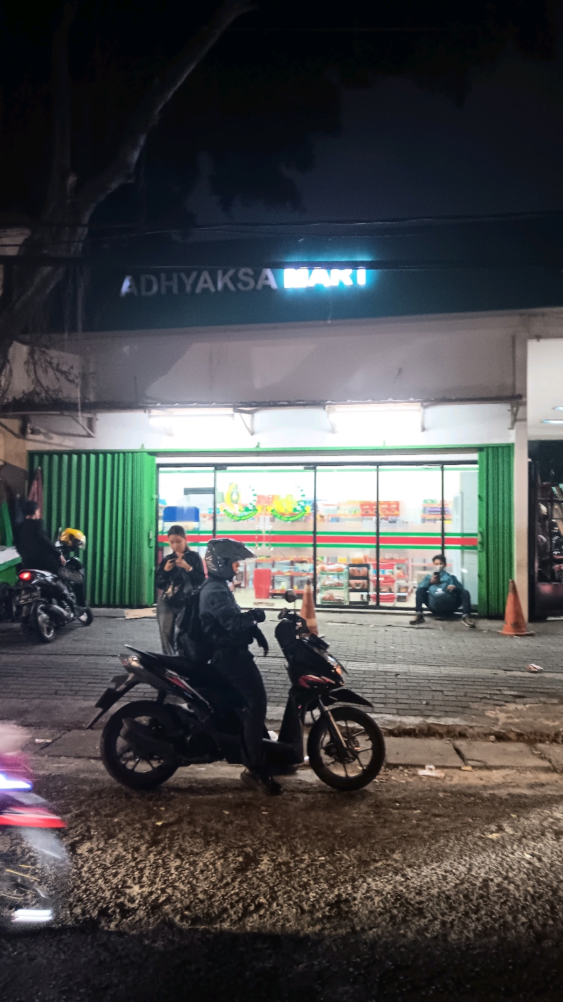 The image depicts a nighttime street scene outside a brightly lit convenience store in South Jakarta, Indonesia. The store's facade is marked with the neon sign "ADHYAKSA MART." A person wearing a black helmet and jacket sits on a black motorcycle in the foreground, facing the store. To the left of the motorcycle, another motorcycle is parked, with a person wearing a black jacket and a yellow helmet standing next to it. Inside the store, the shelves are stocked with various products, visible through the large glass windows. A woman is standing near the store entrance, looking at her phone. Further inside, a person wearing a blue jacket is seated on the floor, also looking at their phone. Several orange traffic cones are visible on the sidewalk. The ground appears to be paved with asphalt and concrete, with some debris scattered around. The overall atmosphere suggests a common urban scene at night.