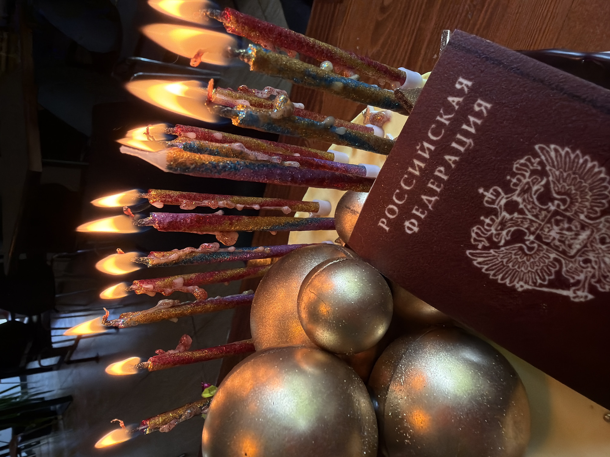 The image shows a cluster of lit candles with decorative glitter. Adjacent to the candles is a dark red Russian passport, printed with the words "РОССИЙСКАЯ ФЕДЕРАЦИЯ" and the Russian coat of arms. Gold-colored spherical objects are also part of the scene. The location is not specified, and there is no basis to conclude this image was taken in the city of Birobidzhan, Russia.