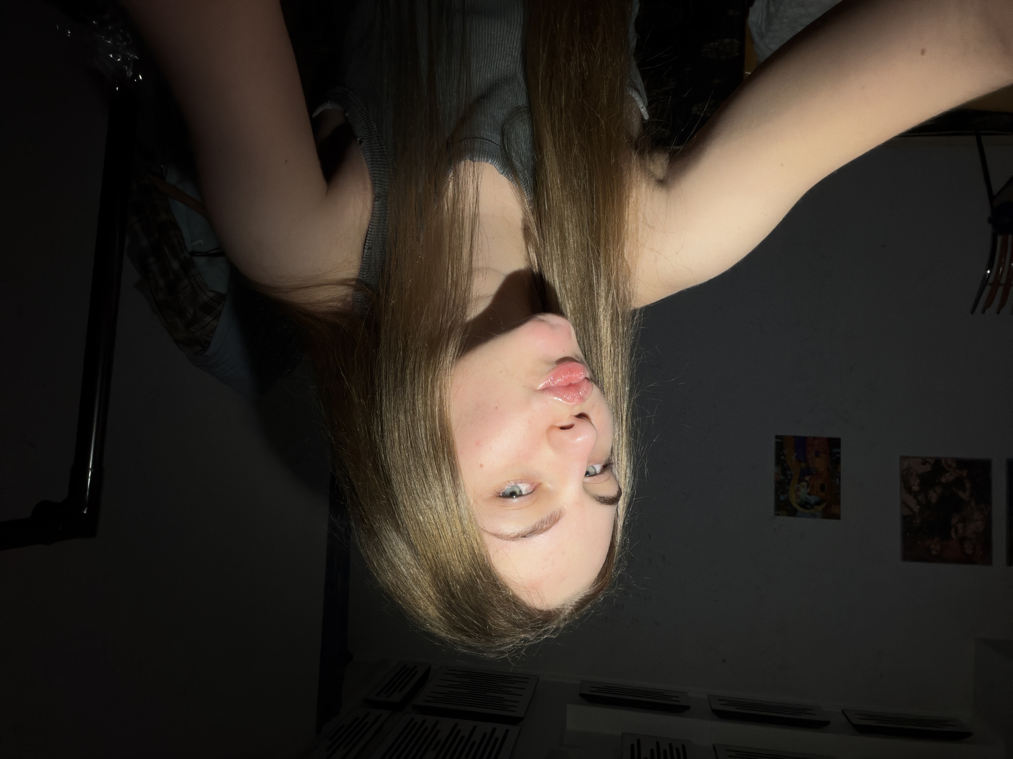 The image is a selfie of a young woman with long, light brown hair and blue eyes. She is wearing a gray tank top and is making a kissing face. She is indoors, with a wall behind her that has a few framed pictures and what appear to be ventilation grilles on the ceiling. The lighting is dim and appears to be coming from the front, casting shadows on the wall and her face. There are no discernible elements in the image that would allow for the precise identification of the city of Dnipro, Ukraine.