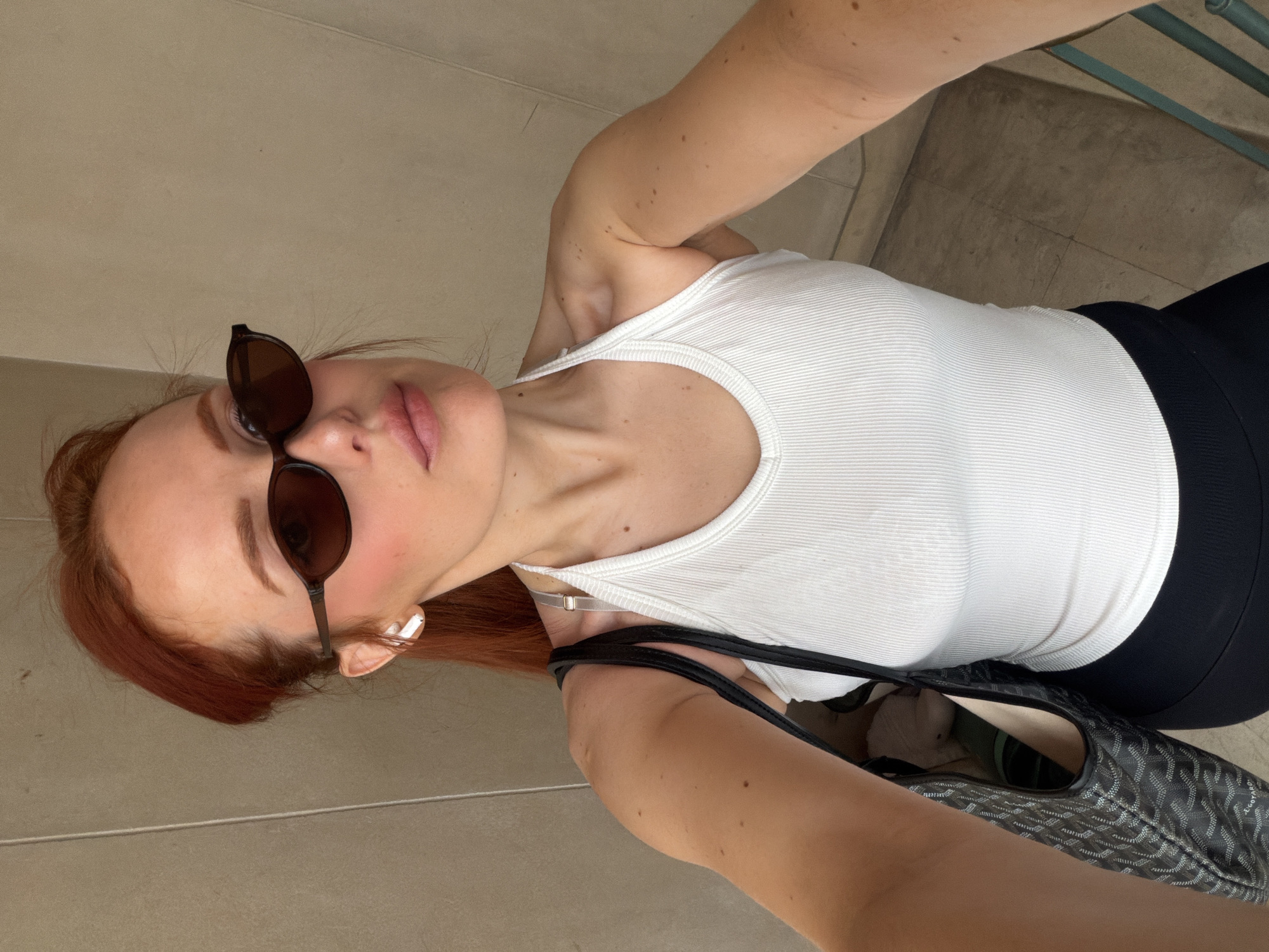 A woman is looking up at the camera, posing for a selfie. She is wearing sunglasses, a white tank top, and black pants. She has red hair and is holding a patterned tote bag. She is also wearing an earbud in her left ear. The background appears to be a neutral tiled surface, potentially a floor or wall. The location is most likely in Paris, France, given the style and the architecture often associated with the city.