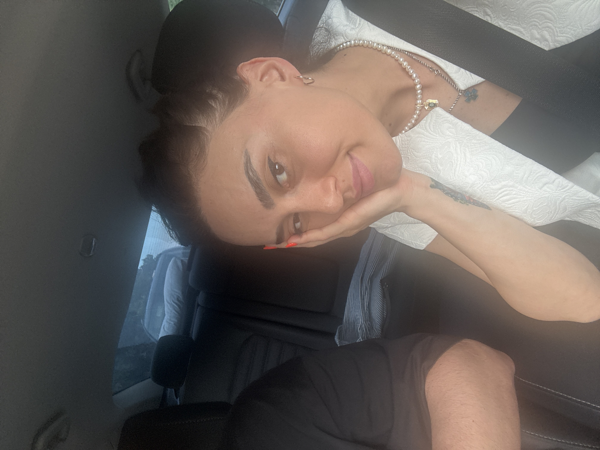 A woman with dark hair pulled back is in the passenger seat of a car, resting her hand on her cheek. She is looking at the camera and smiling slightly. She is wearing a white textured jacket and a pearl necklace with two pendants. A seatbelt is across her chest. The interior of the car is visible, including the headrest and part of the driver's seat. Outside the car, trees and a blue sky are visible through the windows. The location is not determinable from the image.