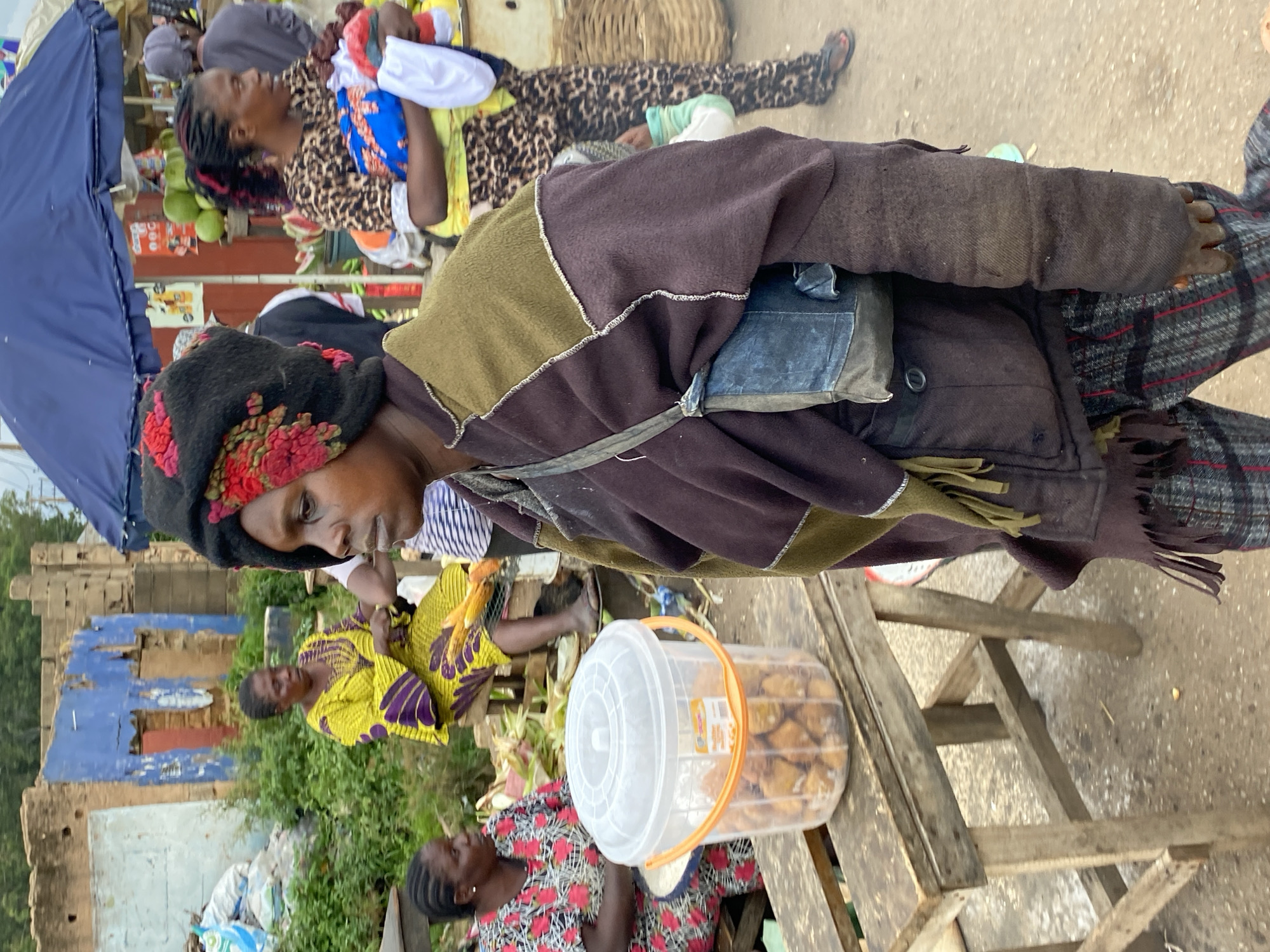 The image depicts an outdoor market scene in Bukuru, Nigeria. A woman in the foreground, wearing a dark, patchwork-like outfit and a black and red patterned headscarf, is the primary subject. She carries a small, dark blue pouch across her chest. To her left, a clear plastic bucket filled with what appears to be fried dough items sits on a rustic wooden table. Behind the main subject, other individuals are visible. One woman in a bright yellow dress with purple accents is seated on a bench, and another woman wearing a leopard-print top and dark pants is holding a baby wrapped in colorful fabric. The background shows a dilapidated building with blue and brown walls and overgrown vegetation, suggesting an urban or semi-urban environment. The ground is a dusty, reddish-brown surface, typical of unpaved areas. The overall impression is of a bustling, albeit somewhat worn, marketplace.