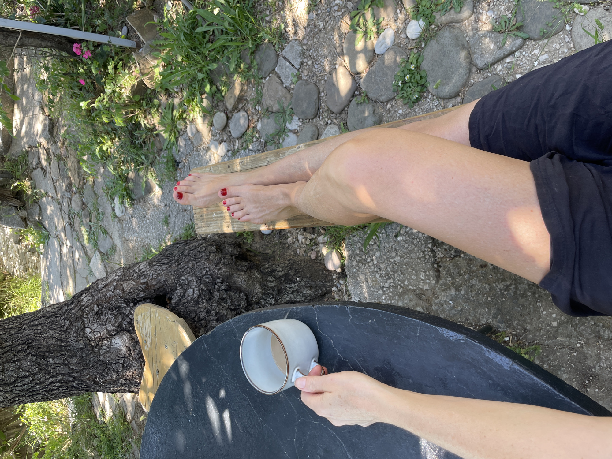 An overhead shot shows a person sitting outdoors in Šušanj, Montenegro, possibly in a garden or patio setting. The person is wearing black shorts and has their legs crossed, resting their feet on a wooden plank. The person's toenails are painted red. To the right of the legs is a round, dark-colored table with a white mug in the lower right corner, being held by a hand. A tree trunk is visible to the left of the table. The ground is covered with stones, dirt, and some vegetation.