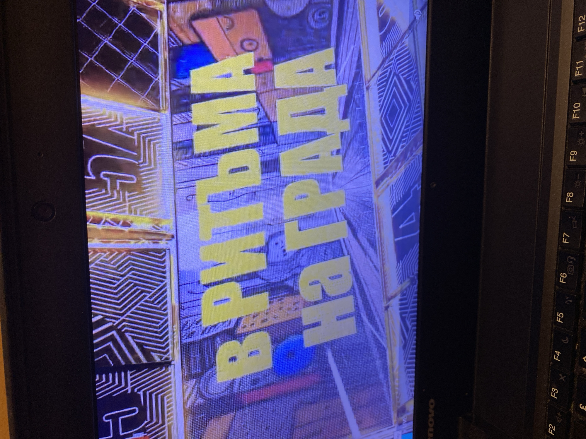 The image shows a close-up of a laptop screen displaying text in Cyrillic. The text appears to be in yellow, bold, block letters and reads "В РИТЪМА НА ГРАДА". The background of the screen is a blurred, colorful pattern with geometric shapes. A portion of the laptop keyboard is visible at the bottom of the image, with function keys labeled F2 through F12. The branding "Lenovo" is visible on the laptop bezel. There is no information in the image to suggest any specific location in Smolyan, Bulgaria.