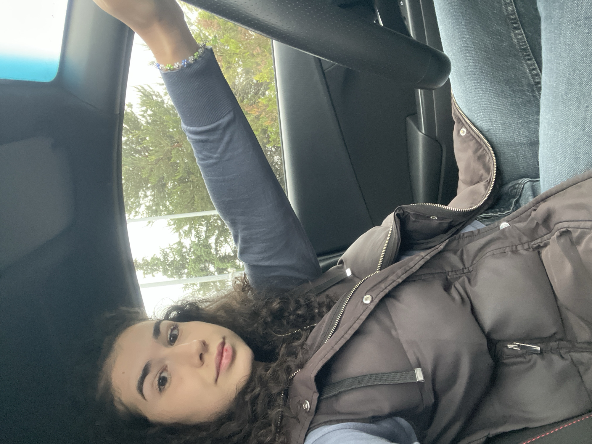 An indoor, eye-level shot, rotated counter-clockwise by 90 degrees, features a young woman lying in what appears to be a car. The woman is positioned on the right side of the rotated frame, with her head towards the bottom. She has curly dark hair, dark eyes, and dark eyebrows, with a neutral expression. She is wearing a dark blue long-sleeved shirt and a dark brown puffer vest with visible zippers and snaps. Her right arm is raised and extended towards the top of the frame, showcasing a delicate, multicolored beaded bracelet (blue, green, white) on her wrist.
The background suggests the interior of a car, with black elements like a dashboard, steering wheel, and a headliner visible. Outside the car window, blurred green foliage from trees is visible under what appears to be an overcast sky, indicating daytime. Light diffuses softly into the car interior. There are no specific external location cues within the image to confirm the provided context of Sofia, Bulgaria. No visible text is present in the image.