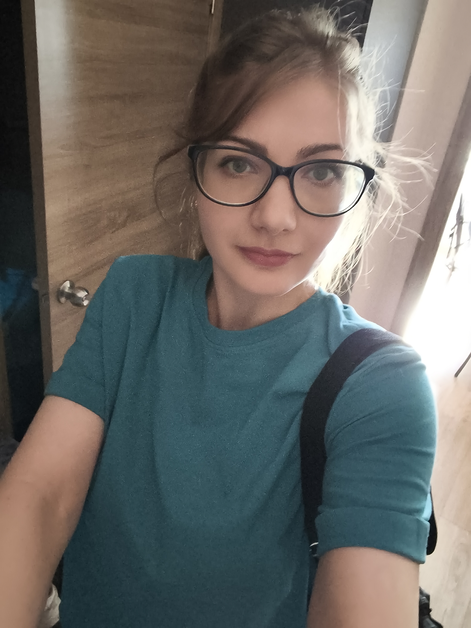 A selfie of a young woman with blonde hair pulled up in a messy bun, wearing glasses and a teal t-shirt, is shown. She has fair skin and light makeup. She is standing indoors, in front of a wooden door and a lightly colored wall. The background suggests a domestic setting. There is no visible indication of the location being in a city.