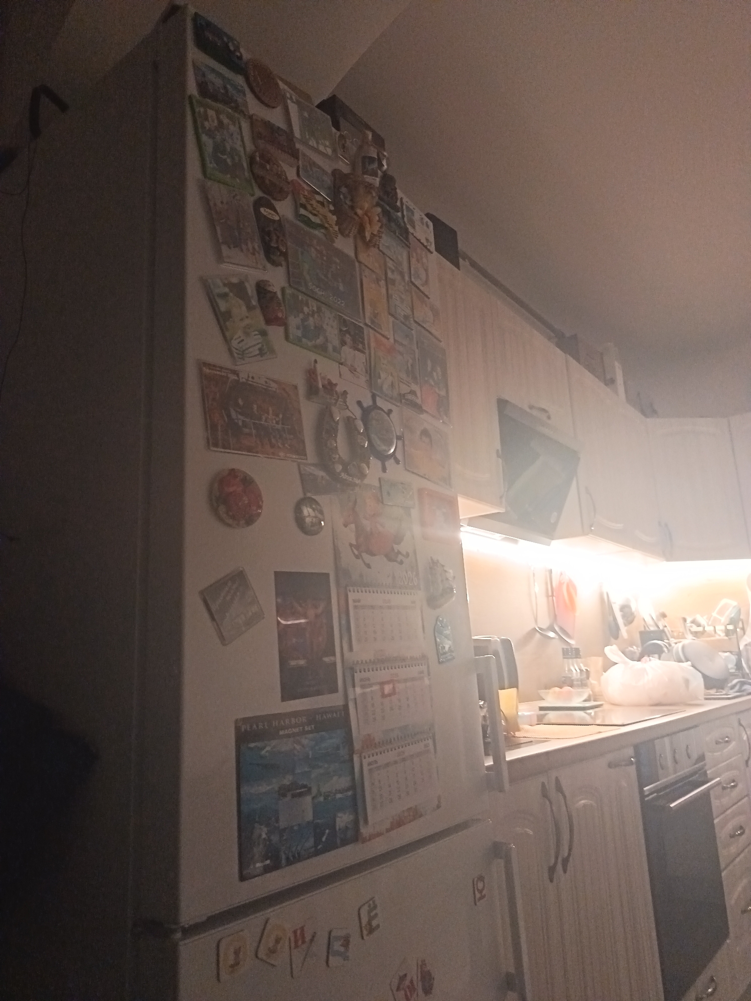This image captures a domestic kitchen scene, likely in the evening or at night, illuminated predominantly by artificial lights. The main subject is a white refrigerator positioned on the left, heavily adorned with a collection of magnets and calendars.
The refrigerator's surface is covered with various souvenir magnets. Visible text includes "SOCHI 2022" on one magnet, directly referencing the provided location context of Sochi, Russia. Another magnet displays "PEARL HARBOR - HAWAII MAGNET SET". Multiple pictorial magnets depict landscapes, illustrations, and decorative items. Two calendars are affixed, with one clearly showing monthly grids for 2023. On the lower part of the refrigerator, magnet letters spell out what appears to be "Любовь" (Lyubov), meaning 'love' in Cyrillic.
To the right of the refrigerator, a kitchen counter extends with white upper and lower cabinets. Under-cabinet strip lighting casts a bright, warm glow across the countertop and the adjacent workspace. On the counter, various kitchen items are visible, including bottles (possibly oil or condiments), a small digital scale with a bowl, a white plastic bag, and some dishes or utensils. A range hood is visible above the stovetop area. No people are present in the scene. The overall ambiance is cozy, characteristic of a lived-in kitchen.