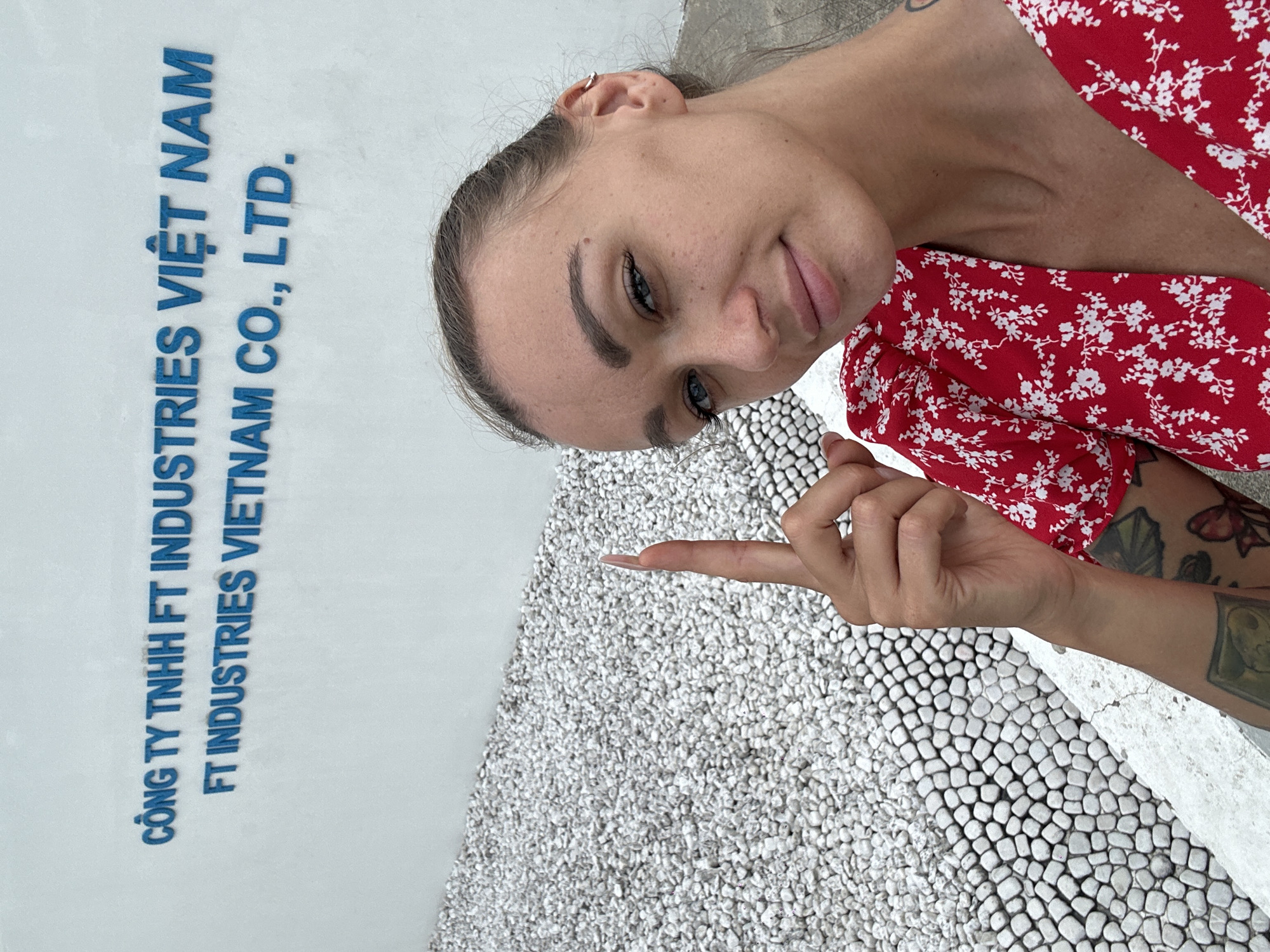 The image shows a woman with a red floral dress and arm tattoos, standing near a wall with blue text that reads "CÔNG TY TNHH FT INDUSTRIES VIỆT NAM FT INDUSTRIES VIETNAM CO., LTD." The woman is pointing her finger at a ground of white and grey pebbles. The text indicates the location is related to a company in Đồng Nai, Vietnam.