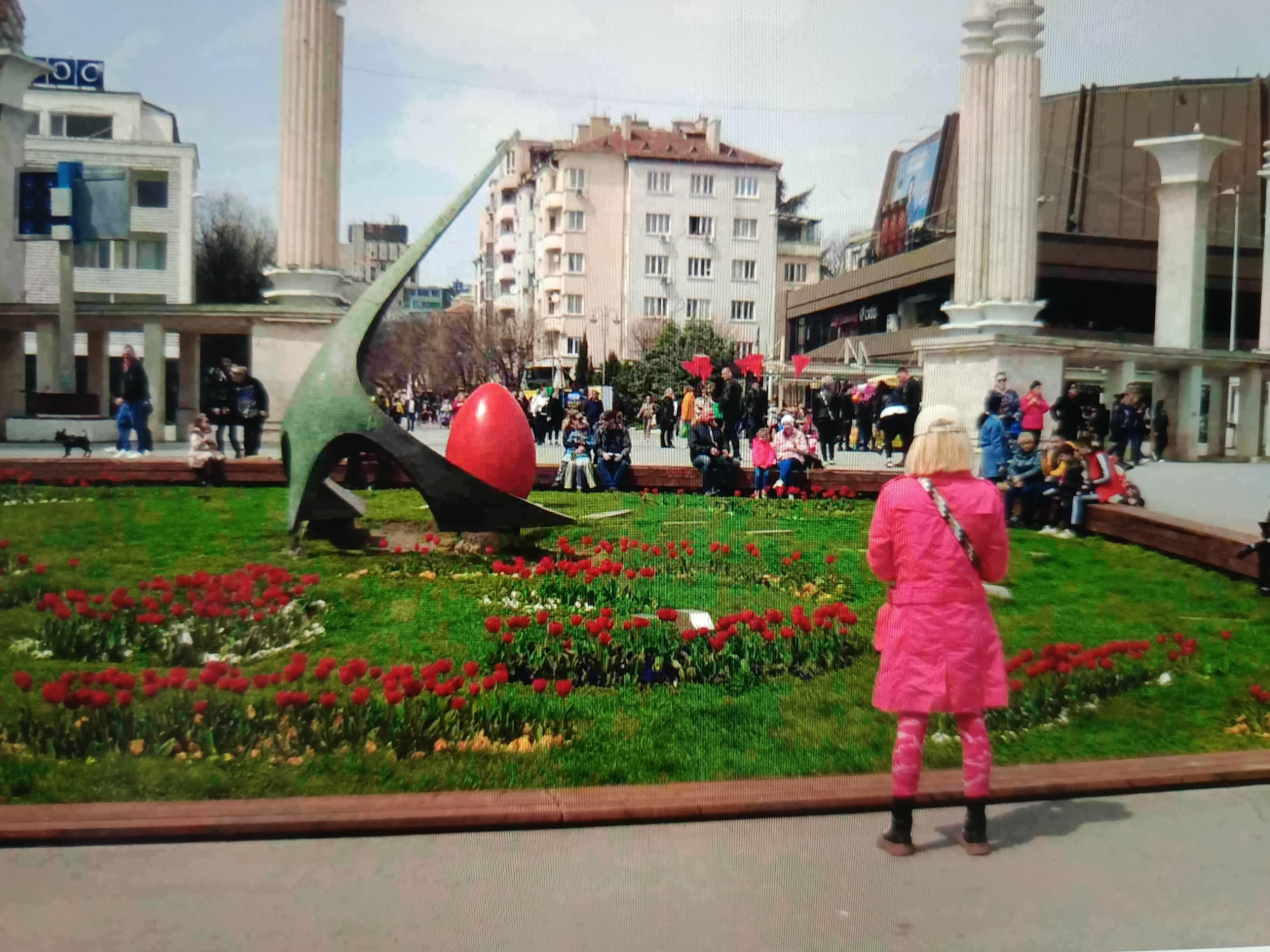 In the city of Varna, Bulgaria, a large red egg sculpture is displayed prominently in a public square, likely as part of an Easter exhibition or celebration. The sculpture is situated within a manicured flower bed filled with vibrant red tulips and other greenery. In the background, a group of people are gathered, some seated on a low wall and others standing, observing the display. Several children are also present. Behind the plaza, urban architecture is visible, including modern buildings and columns from what appears to be a more classical structure. A woman in a bright pink coat and patterned leggings is facing away from the camera, looking towards the egg sculpture. The overall scene suggests a festive outdoor gathering on a bright day.
