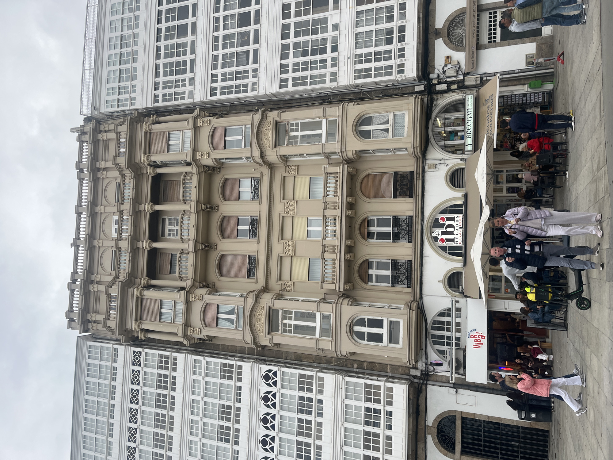 This media file depicts a bustling street scene in A Coruña, Spain, characterized by distinctive architecture and a lively pedestrian environment under an overcast sky.
The central feature of the image is a multi-story building with ornate, classical architecture, rendered in a light beige color. It boasts decorative balconies with balustrades, columns, pilasters, and arched window frames. This central structure is flanked by taller buildings on both sides, which showcase the iconic "galerías" – white glazed enclosed balconies that are a hallmark of A Coruña's architecture. These buildings feature numerous grid-patterned windows.
At street level, the scene is active with commercial establishments. On the right, an outdoor seating area with white umbrellas, tables, and chairs suggests a cafe or restaurant. A shop with a broad awning displays the text "Regalos SOUVENIRS." Another storefront shows "ACAI CLUB VIBE!" and "IO HELADOS Desde 1962." Pedestrians are seen walking along the pavement, some stopping to socialize or patronize the businesses. People are dressed in casual clothing.
Notable details include the uniformly gray, cloudy sky, indicating an overcast day. The architecture, especially the "galerías," provides a strong geographical cue for A Coruña.
Visible text includes:
* "AGAI"
* "IO HELADOS Desde 1962"
* "ACAI CLUB VIBE!"
* "BREOGAN"
* "DOGAMOS"
* "Regalos SOUVENIRS"
* "Estrella Galicia"
* "www.restaurantenor"