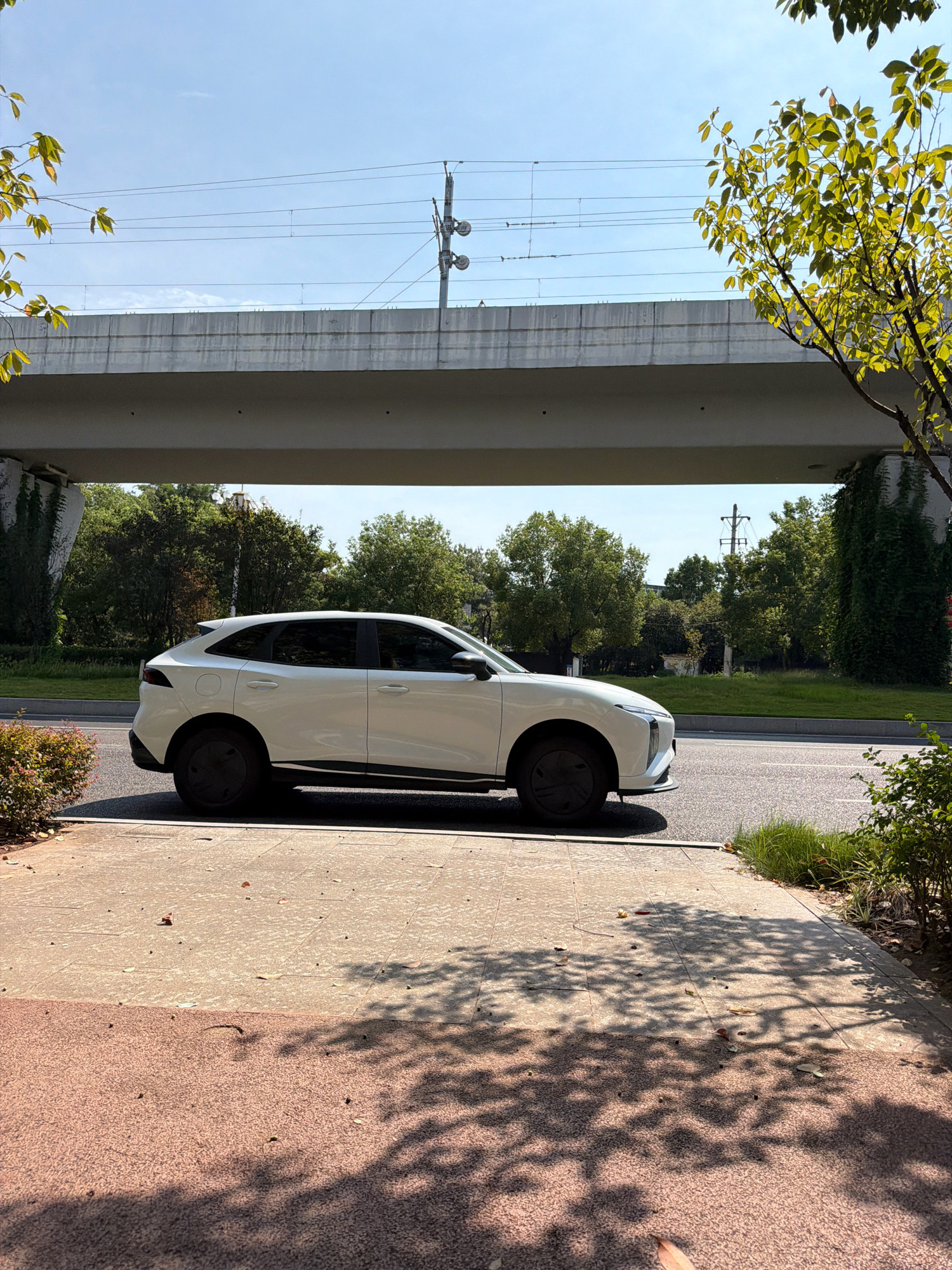 A white SUV is parked on the side of a road, under an elevated concrete structure, likely a bridge or overpass. Overhead, power lines run parallel to the structure, supported by utility poles. The area around the road is lined with greenery, including trees and bushes. The ground in the foreground is paved with large, light-colored tiles, with shadows from overhead foliage creating a dappled pattern. The scene appears to be in an urban environment, possibly in Jiangjin, China, given the context of the query. The sky is clear and blue, suggesting a sunny day. There are no people visible in the image.