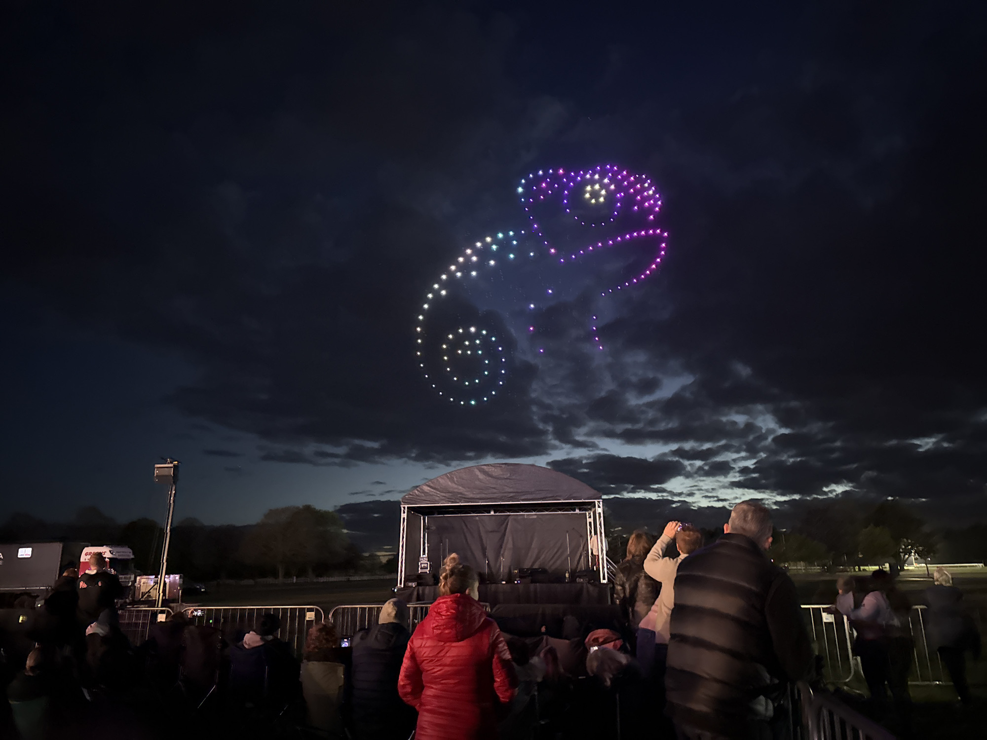 A crowd of people watches a drone show in South Norfolk, United Kingdom at night. The drones create the shape of a chameleon in the sky with colorful lights. In front of the crowd is a stage. A truck with its crane-lift is seen to the left of the stage. The sky is dark with clouds.