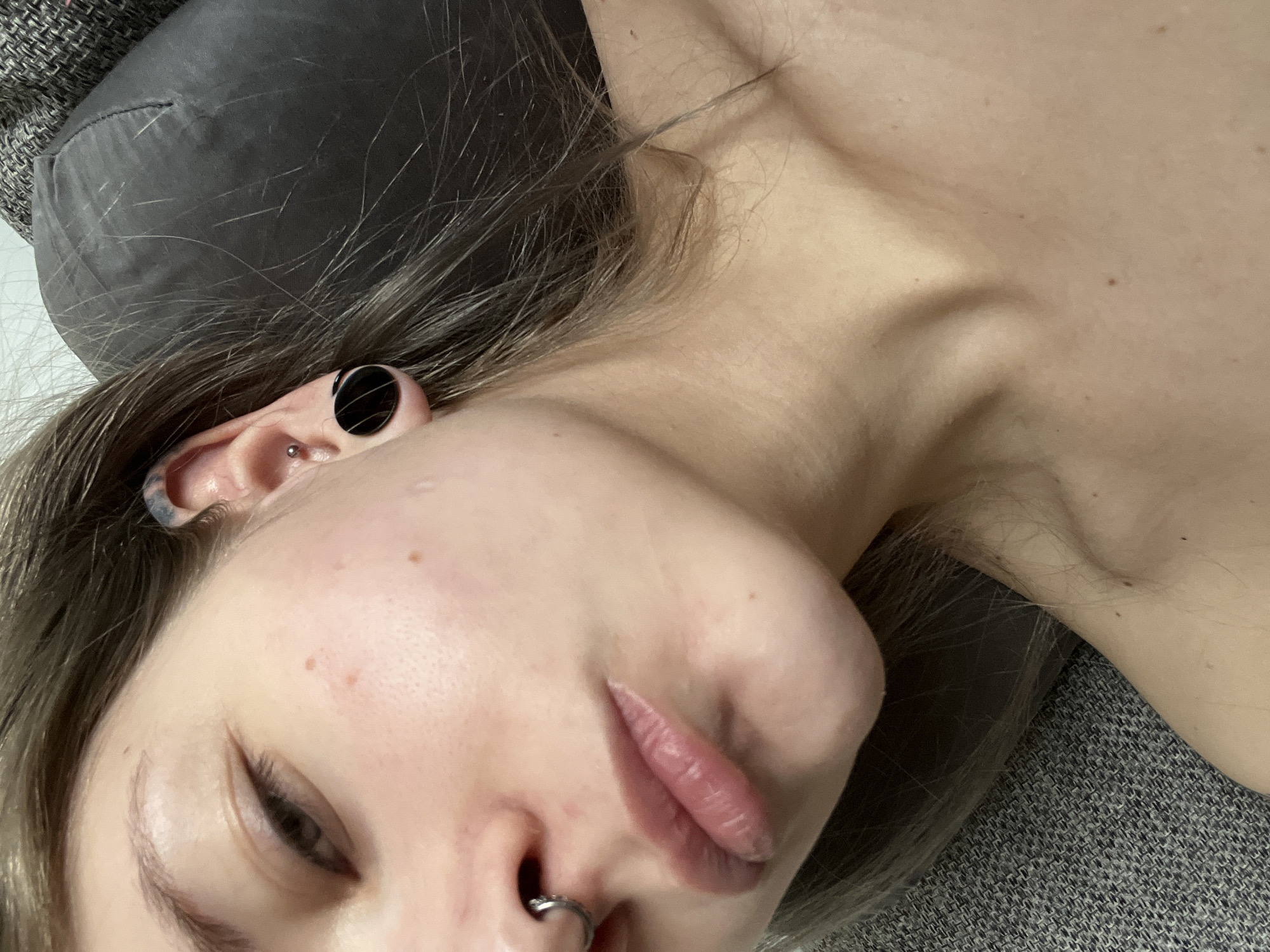 A close-up, angled view of a person with pale skin lying down. They have brown hair with lighter streaks, a piercing in their nose, multiple piercings in their left ear including a large gauge, and closed eyes. The person is resting on a grey textured surface that appears to be a couch or similar upholstered furniture. There is a grey cushion or headrest behind their head. The setting is a domestic or indoor environment. There is no way to determine the location is Khimki, Russia from the image.