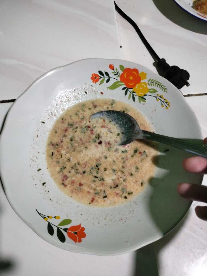 A close-up, slightly overhead shot captures a white ceramic plate containing a liquid dish. The plate features a decorative floral pattern along its rim, with vibrant orange, yellow, and green flowers and leaves. Inside the plate is a creamy, light beige liquid abundantly flecked with small green pieces, likely chopped herbs, and tiny red specks, possibly chili flakes or other spices. A silver-colored spoon, with a partial view of its green handle, rests in the liquid, its bowl submerged.
A person's hand is partially visible on the right side of the frame, positioned as if holding or reaching for the plate. The plate rests on a light-colored surface, possibly a tiled floor or tabletop, which shows faint dark lines between the tiles. In the upper right corner, a black electrical cord with a two-pronged plug is visible. The lighting is artificial, casting noticeable shadows, suggesting the photograph was taken indoors, likely during the evening or night in Jasinga, Indonesia. There is no visible text.