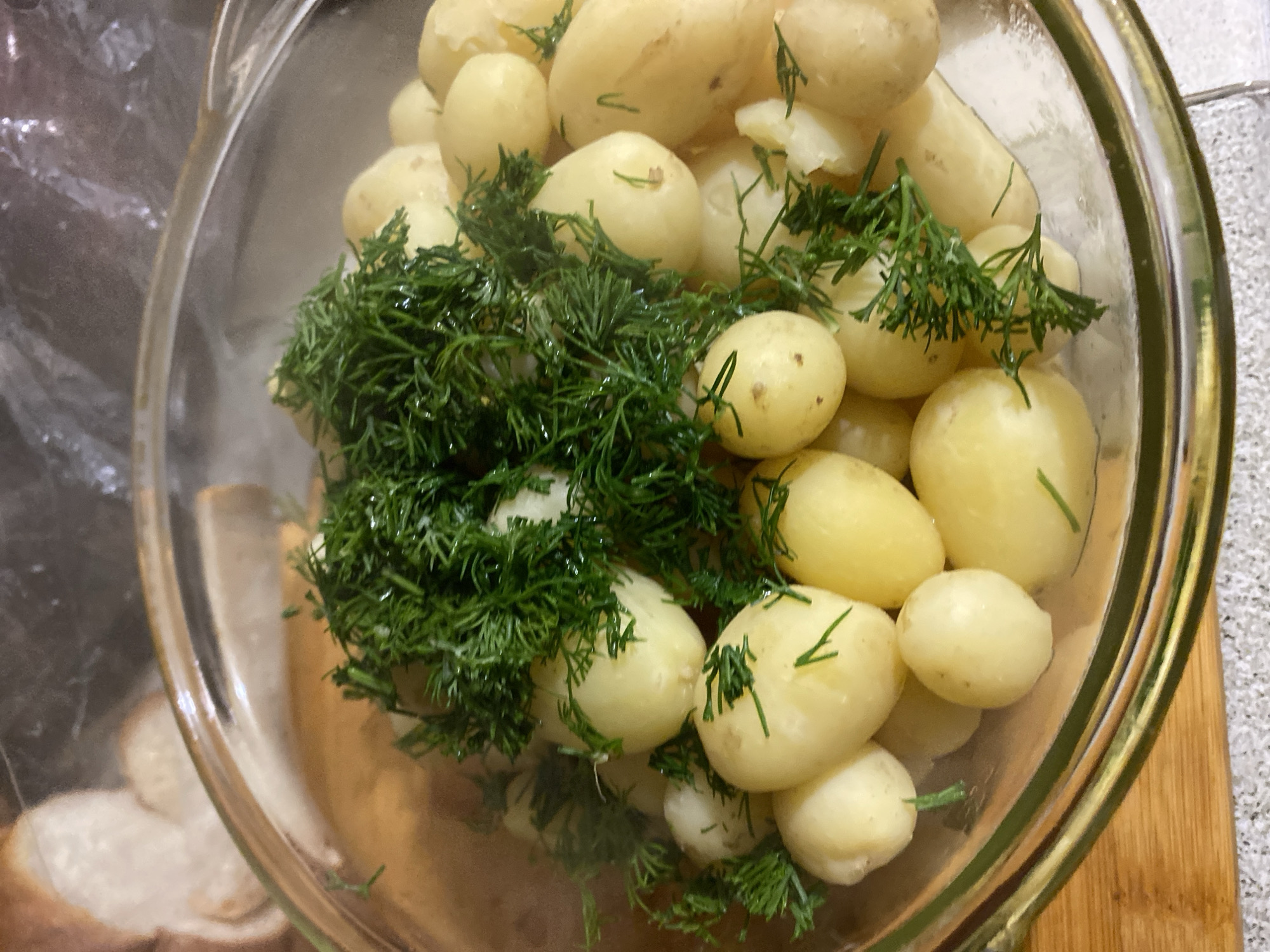 A close-up, overhead shot showcases a clear glass bowl filled with small, pale yellow, boiled potatoes. The potatoes are clustered together, some still partially submerged in a liquid that is not fully visible. Fresh dill, with its delicate, feathery fronds, is generously scattered amongst the potatoes, some sprigs appearing damp. The dill is a vibrant green and contrasts with the lighter color of the potatoes. The surface of the potatoes and dill appears wet, suggesting they may have been recently prepared or are being marinated.
The bowl itself has a slightly metallic rim, possibly silver or a similar reflective material, adding a subtle sheen to the image. The background is partially visible and appears to include a textured surface, possibly a countertop or table, with hints of a wooden cutting board to the right. A blurred plastic wrap or bag is visible on the left side of the frame, partially obscuring some of the bowl's exterior. The lighting is relatively bright, highlighting the textures and colors of the food. There is no visible text in the image. The scene depicts the preparation of a simple dish, likely a side dish or salad, emphasizing fresh ingredients. The location context, Маринопіль, Ukraine, suggests this dish might be part of a meal or cooking process within that region.