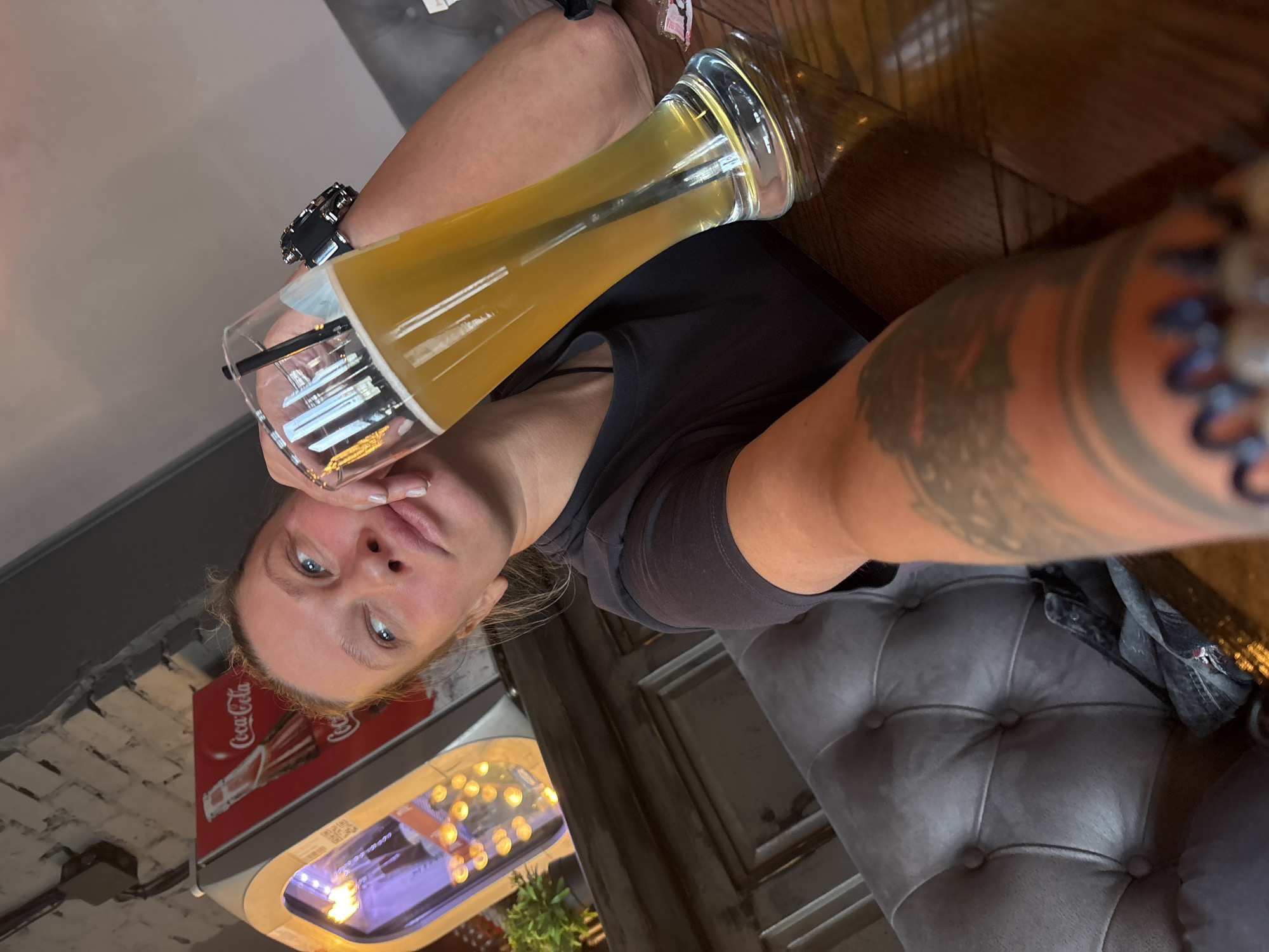 A selfie taken indoors in Perm, Russia shows a woman with fair skin and blonde hair holding a tall, narrow glass filled with a pale yellow liquid, possibly beer. She is wearing a dark-colored t-shirt and a large watch on her left wrist. Her right arm, extending towards the camera, has prominent tattoos. She is seated on a tufted grey chair. Behind her, a Coca-Cola advertisement is visible next to a lighted display containing small, round objects. The background also shows a brick wall and wooden surfaces.