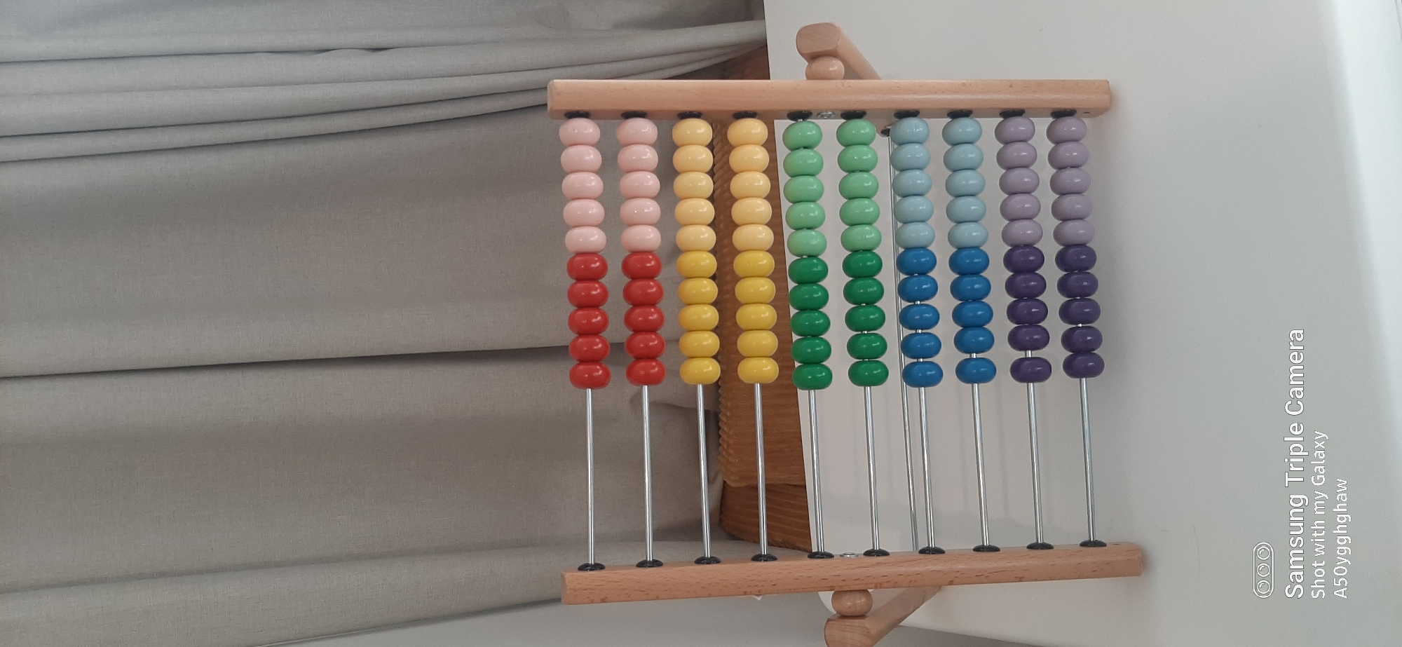 An abacus with wooden frame and ten rows of beads rests on a white surface in front of a beige curtain. The beads are colored red, pink, yellow, green, blue, and purple, arranged in alternating colors on each row. The abacus is positioned centrally in the frame. The environment appears to be indoors, with soft lighting. There is no indication of the location being Dubai, United Arab Emirates.