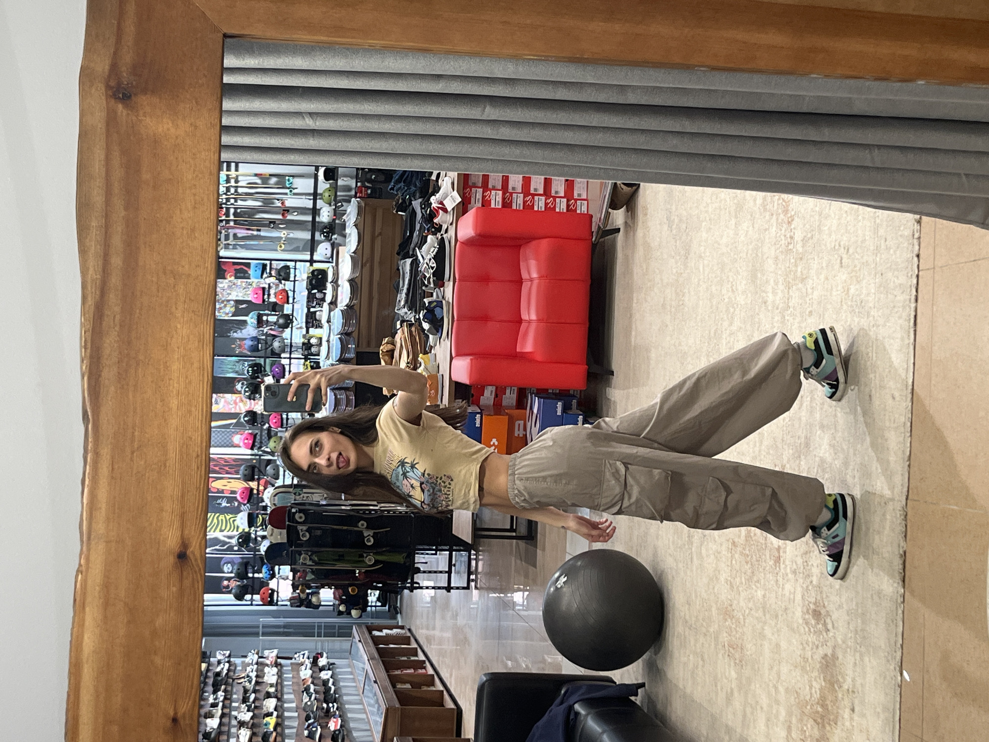 A woman is taking a selfie in a full-length mirror. The mirror has a wooden frame. The reflection reveals a retail store setting with various merchandise displays including skateboards, helmets, shoes, and red padded furniture. The woman is wearing a short-sleeved shirt, loose pants, and sneakers. In the foreground of the reflection there is a black exercise ball and a dark chair. The store is possibly located in Krasnoyarsk, Russia, but there is no explicit visual indicator of this in the image.