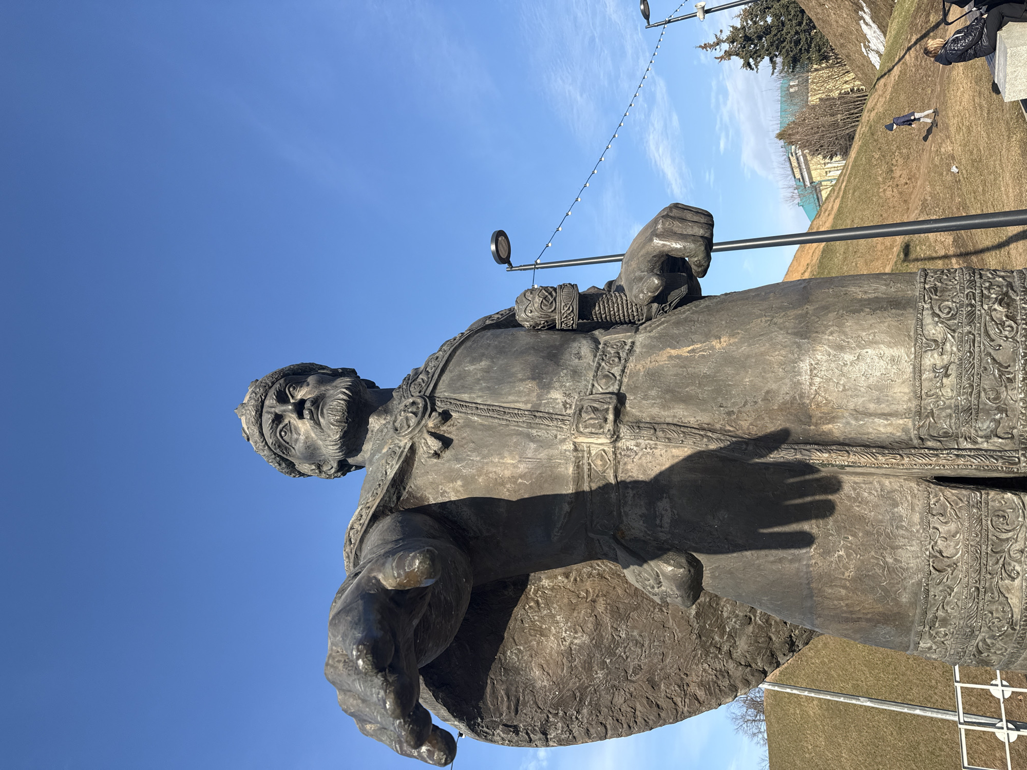 A low-angle, wide shot captures the bronze monument to Yuri Dolgoruky in the Dmitrov Kremlin, Dmitrov, Russia, on a clear, sunny day. The large statue, occupying the central and left foreground, depicts a bearded male figure wearing a crown and ornate robes, with his right arm extended forward, pointing. His left hand grasps the hilt of a weapon. The statue casts a distinct shadow on its lower body.
The background features a bright blue sky with sparse, wispy clouds. To the right and behind the statue, grassy earthen embankments rise, showing patches of dry, brown grass mixed with remnants of snow in shaded areas. Scattered leafless trees are visible on the slopes and in the distance. Two dark grey modern streetlights with circular luminaires are present; one stands directly behind the statue's left arm, and the other is further to the right. A string of small, white spherical lights is suspended horizontally between these two streetlights.
Two individuals are visible on the right side of the frame, against the grassy slope. One person, appearing to be a child dressed in a dark jacket and hat, walks uphill on the dry grass. Another individual, wearing a dark quilted jacket, is seated on a light-colored stone bench in the lower right corner, partially obscured by the statue's base, next to what appears to be a dark-colored stroller. A building with a turquoise roof is discernible in the far background, behind the slopes.
