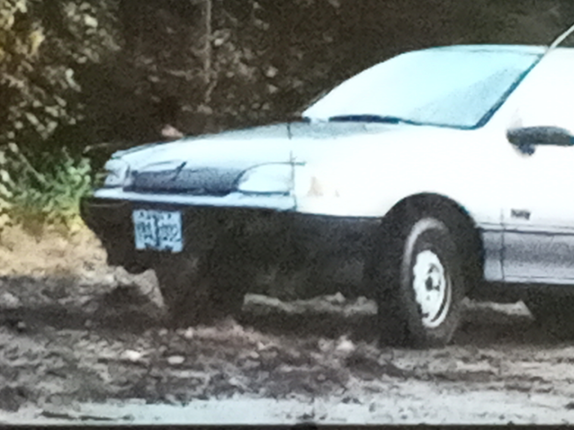 The image shows a white or light-colored compact car, possibly a sedan, parked on a dirt or gravel surface. The car's front end is visible, including the headlight, bumper, and license plate. The background features foliage, suggesting a somewhat rural or wooded area. It is difficult to ascertain whether the car is in the city of Gwong, Nigeria.