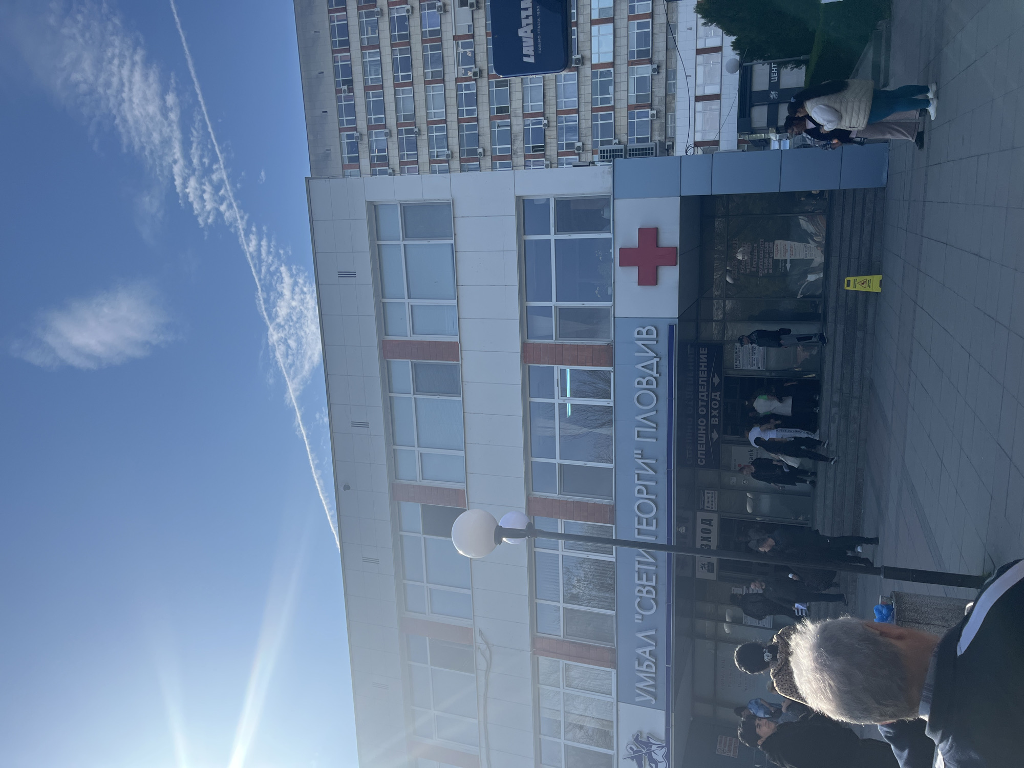 The image shows the entrance of the "Saint George" University Multiprofile Hospital for Active Treatment (УМБАЛ "Свети Георги") in Plovdiv, Bulgaria. The building is multi-storied and modern, featuring a combination of white and gray panels with red accents. The hospital's name is prominently displayed in Bulgarian Cyrillic above the entrance. A red cross is visible on the facade. People are present at the entrance, some appearing to be entering or exiting the hospital. There are signs indicating "emergency department entrance" and "entrance." A "Lavazza" coffee advertisement is seen on an adjacent building. The sky is visible with some cloud formations.