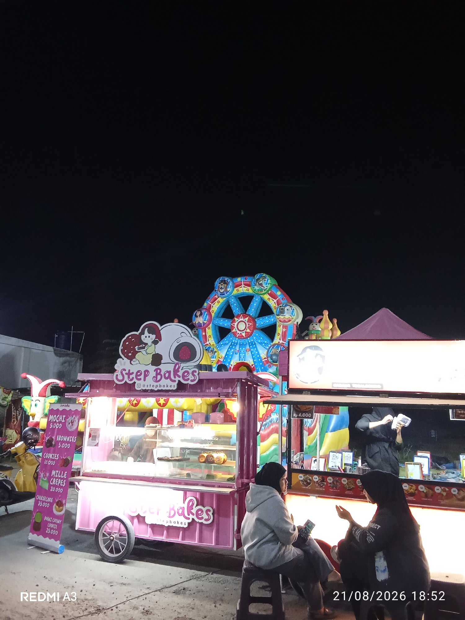 A night scene at an outdoor food stall, identified as "Step Bakes" in Gentan, Indonesia. A pink food cart with a Ferris wheel-like decoration above it is the main subject. The cart displays a menu with items like Mochi, Brownies, Cookies, Mille Crepes, and Cheesecake. Two individuals wearing hijabs are seated on stools in front of the stall, one on the left in a grey hoodie and the other on the right in a black hoodie. The person on the left is looking at a mobile phone, while the person on the right appears to be talking to them. A scooter is parked to the left of the food cart. The background shows a dark night sky with some structures and lights. A sign at the bottom left of the image reads "REDMI A3", and a timestamp at the bottom right reads "21/08/2026 18:52".