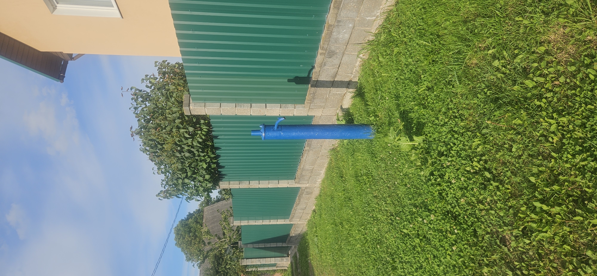 The image depicts a sunny outdoor scene. In the foreground, a patch of green grass with some clover and weeds is visible. A blue water pump stands prominently in the grass, casting a shadow to its right. Behind the pump is a green corrugated metal fence, supported by concrete pillars and a concrete block base. A lush green tree with dense foliage is situated behind the fence, partially obscuring a building with a beige wall and a green roof visible in the upper right quadrant. The sky is blue with scattered white clouds. The scene appears to be in a residential area. While the image does not explicitly state the location, this type of fencing, water pump, and building style are common in various parts of Belarus, including Vitsebsk.