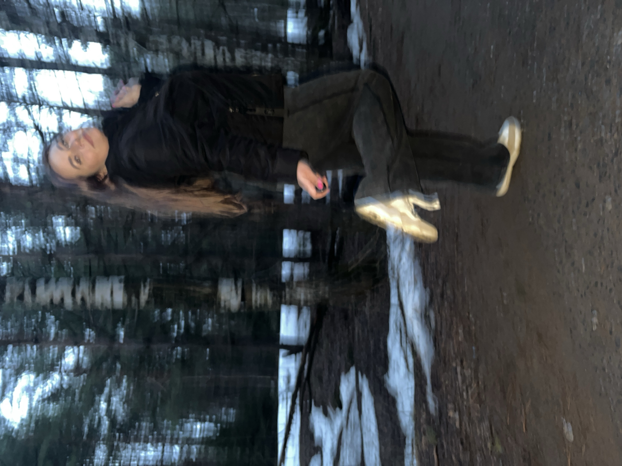 The image shows a woman lying horizontally on a path in a wooded area. The woman is wearing a dark jacket and pants, and light-colored shoes. The background is blurred, with trees on one side and a path with some snow or ice on the other. The location appears to be outdoors, possibly in a park or forest. There is no definitive evidence to confirm the location in Chaikovsky, Russia.