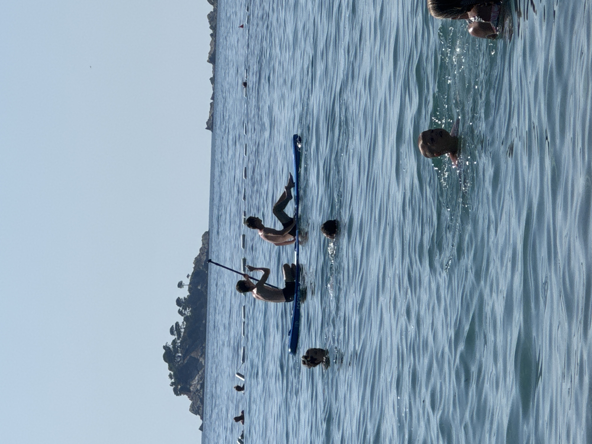 In the waters off the coast of Đurovići, Montenegro, two young males are seen on a paddleboard. The paddleboarder on the left is seated and holds a paddle, while the one on the right is lying down. Several other individuals are in the water, swimming or wading. In the background, a small, vegetated island rises from the sea, and the sky is clear and blue. A marker buoy with a red top is visible further in the distance. The water is calm with gentle ripples, reflecting the bright daylight.