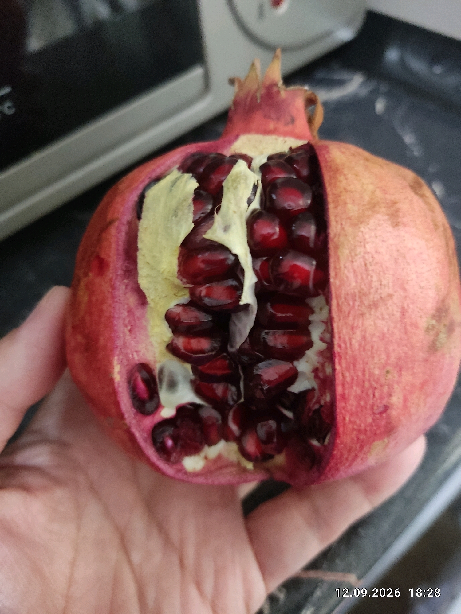 A partially opened pomegranate is held in a person's left hand. The pomegranate is split to reveal its arils, which are a deep red color. The rind of the pomegranate is a reddish-pink. In the background, a blurry metallic appliance is visible. A timestamp in the lower right corner of the image reads "12.09.2026 18:28". There is no discernible information to indicate the location in Bishkek City, Kyrgyzstan.