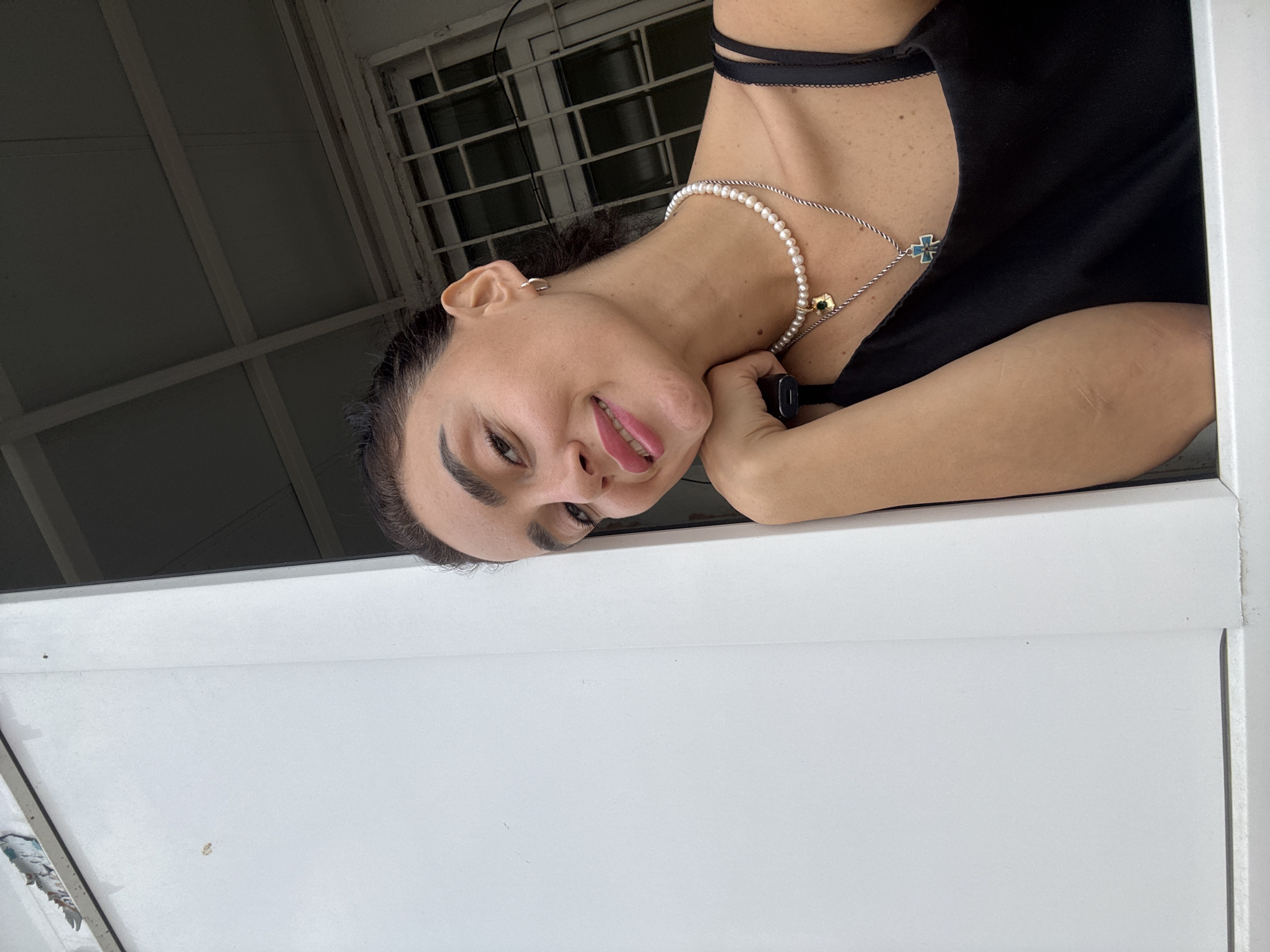 A young woman with dark hair pulled back and prominent eyebrows smiles at the camera. She is wearing a black tank top and a pearl necklace with two pendants: one shaped like a four-leaf clover with a green stone, and another in the shape of a cross with blue and white details. Her left arm rests on her chin, and her right arm is visible at her side. She is leaning against a white surface, possibly a wall or a partition. In the background, a window with a metal grate is visible, suggesting an indoor or semi-outdoor setting. The presence of a ceiling with exposed beams hints at a structure like a porch or a covered outdoor area. The overall impression is a casual, candid portrait.
The image does not contain sufficient information to pinpoint its exact location within Tbilisi, Georgia. However, the architectural elements visible, such as the metal-grated window and the exposed ceiling beams, are common in various residential and commercial buildings across the city. The style of the window grate could suggest older architecture often found in districts like the Old Town or Vera.
