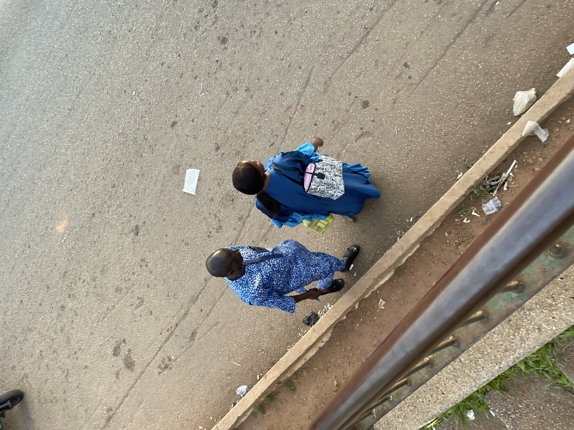 Two individuals, a man and a woman, are walking on a paved surface, likely a road or sidewalk, in Gura Top, Nigeria. The man is on the left, wearing a blue patterned traditional African garment. The woman is to his right, dressed in a long blue garment and carrying a patterned handbag. Scattered debris, including white paper or plastic, is visible on the ground around them. A metal railing is present in the foreground.