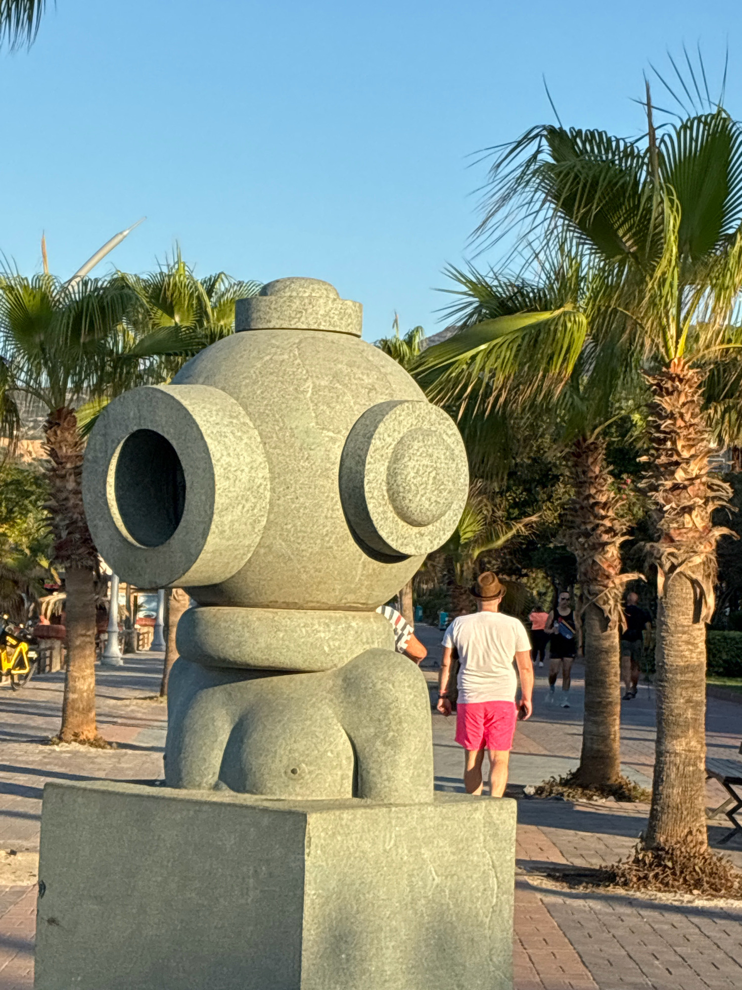The image shows a large, abstract stone sculpture that resembles a diver's helmet. The sculpture is positioned outdoors in a paved area lined with palm trees. In the background, several people are walking. One man in the foreground is wearing a white t-shirt and bright pink shorts, and a straw hat. The scene appears to be in Alanya, Turkey, with palm trees and a clear blue sky suggesting a warm climate.