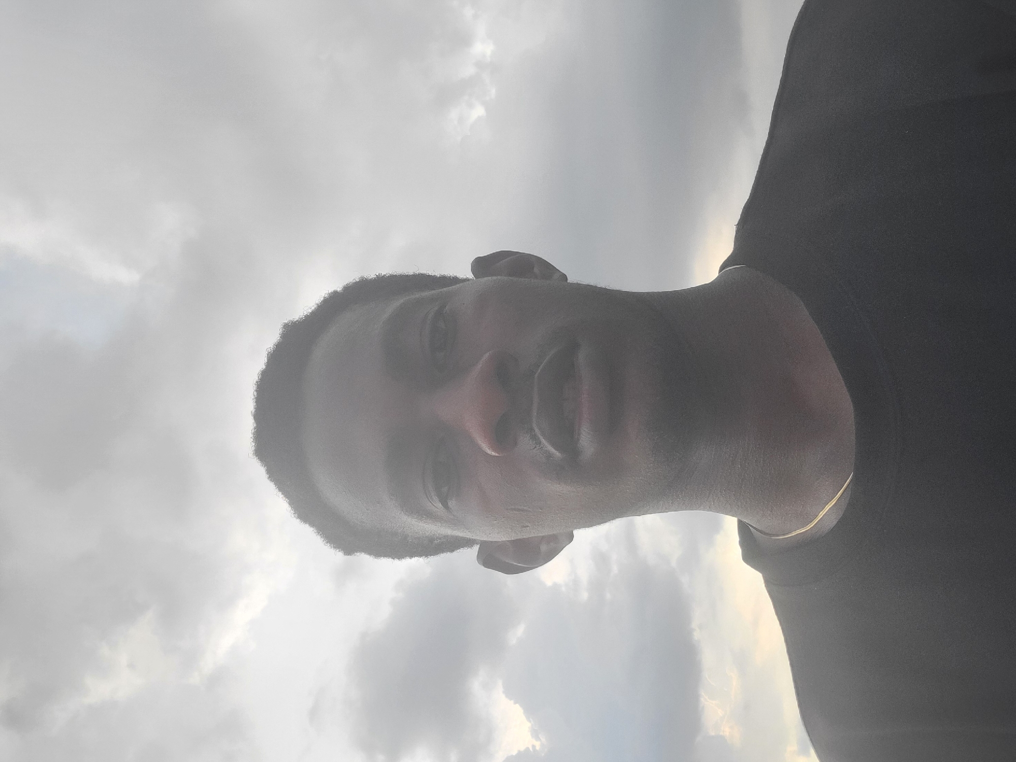 The image depicts a dark-skinned adult male positioned centrally against an overcast sky in Egbagu, Nigeria. He is looking directly into the camera, with his head and shoulders occupying the lower portion of the frame. He has short, dark, curly hair and is wearing a dark, crew-neck t-shirt. A thin, gold-colored chain is visible around his neck. His face is partially shadowed, revealing discernible eyes, nose, and slightly parted lips. The background is an expansive, cloudy sky composed of grey and white clouds, with areas of diffused light present.
