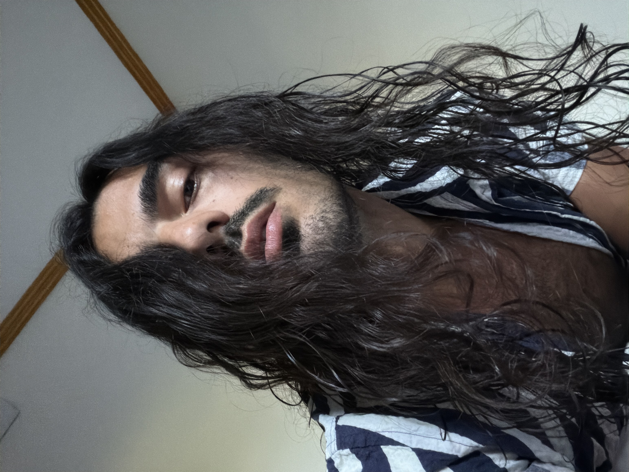 A close-up, angled portrait features a person with long, dark, wavy hair cascading across their face and shoulders. The subject, likely male, has olive-toned skin, thick dark eyebrows, and a short beard or prominent stubble. Their lips are full, and one eye is visible, with a direct, somewhat intense gaze. The hair appears slightly damp or styled to look wet, reflecting some light. They are wearing a dark blue and white striped shirt, which seems to be open at the collar, revealing a glimpse of their chest.
The photograph is taken from a low angle, with the person's head dramatically tilted, giving the image a dynamic and intimate feel. The background is minimalist, consisting of a plain, light-colored wall or ceiling, with a narrow, light-brown strip running diagonally in the upper left corner. The lighting is soft and even, highlighting the contours of the face and the texture of the hair. The setting is likely indoors, possibly in North Kuta, Indonesia. There is no visible text in the image.