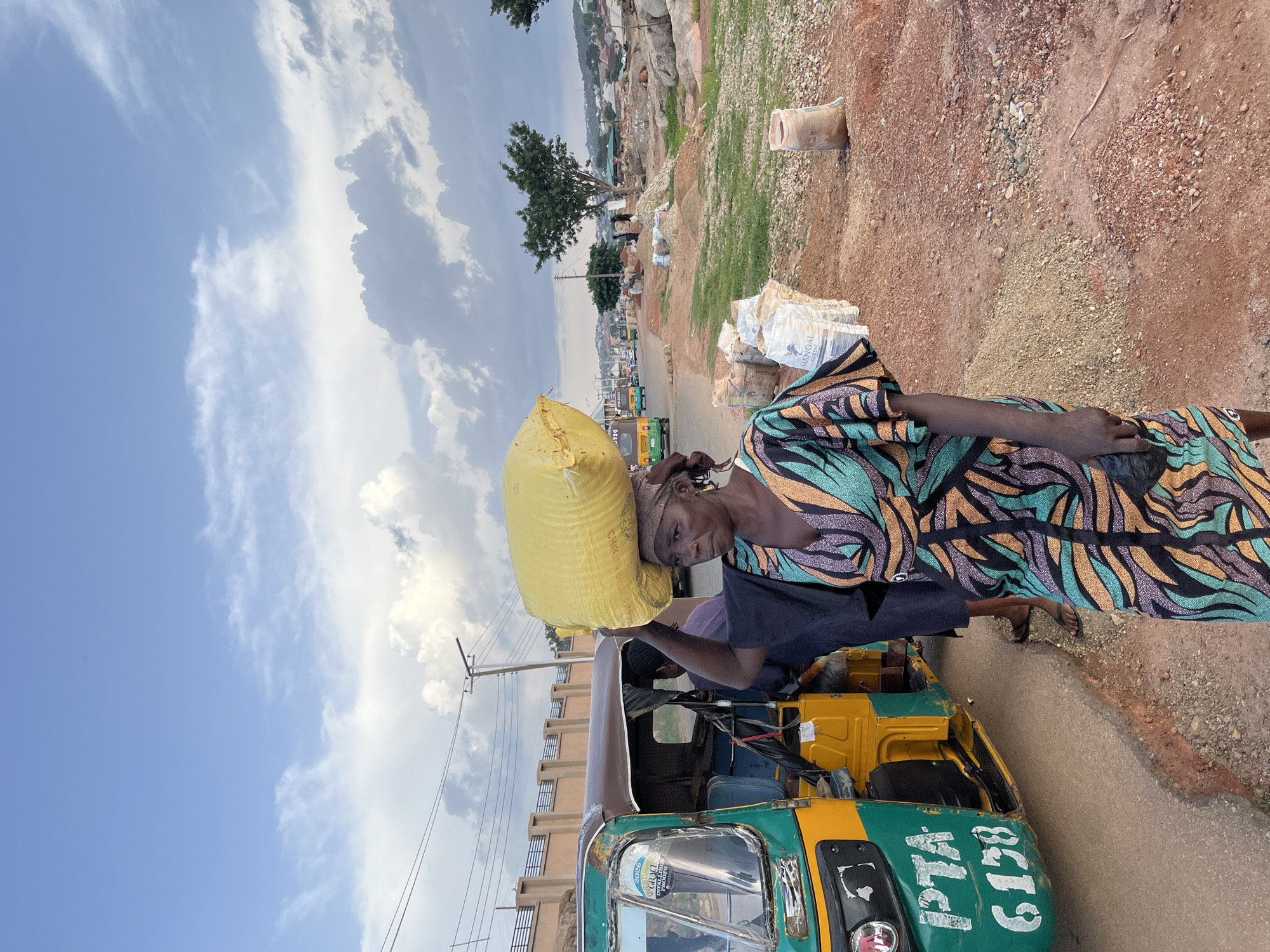 A woman carries a large yellow bag of goods on her head while walking beside a green and yellow auto rickshaw. She is wearing a colorful dress and a headscarf. The scene appears to be a roadside market in Jos, Nigeria, with piles of sand and cement bags visible in the background. The sky is cloudy, and there are buildings and trees in the distance. The auto rickshaw has license plates visible. The ground is dirt and gravel.