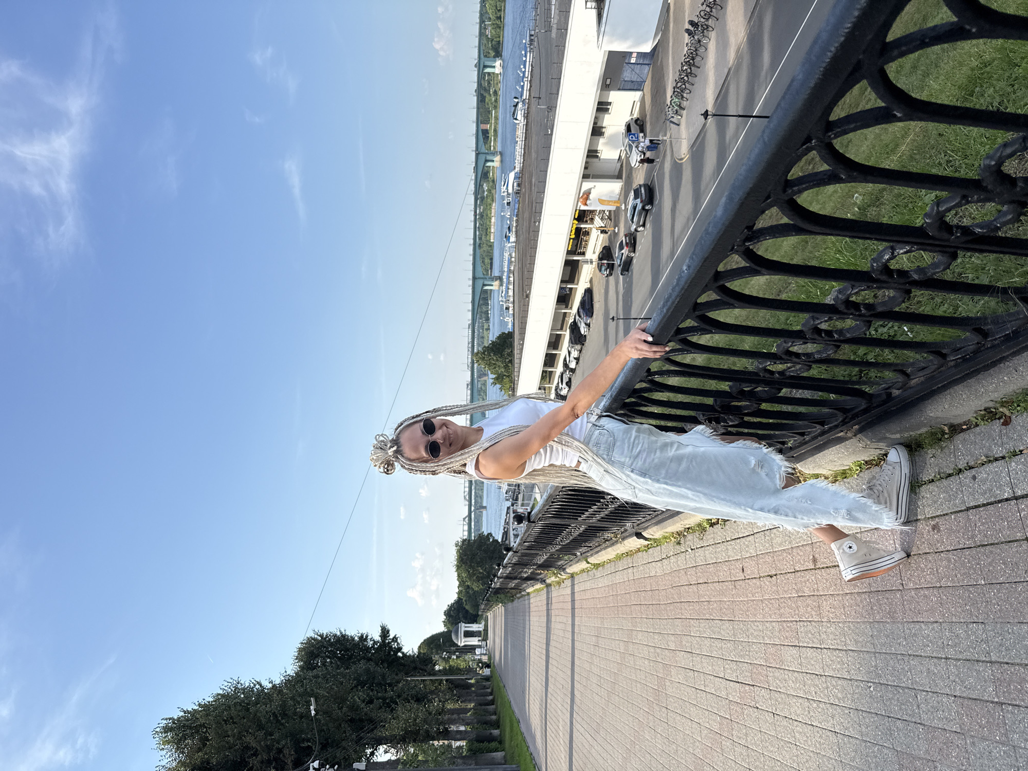 A woman with blonde braided hair and sunglasses is leaning on a black metal railing, smiling at the camera. She is wearing a white tank top and light blue distressed jeans. She is standing on a paved walkway, which is lined with trees on the left side and a body of water with a bridge in the background. The walkway appears to be a promenade or embankment. In the background, there is a body of water, a bridge, a building with parked cars, and a line of bicycles. The sky is blue with some scattered clouds. The location is identified as Rusanovo, Russia.
